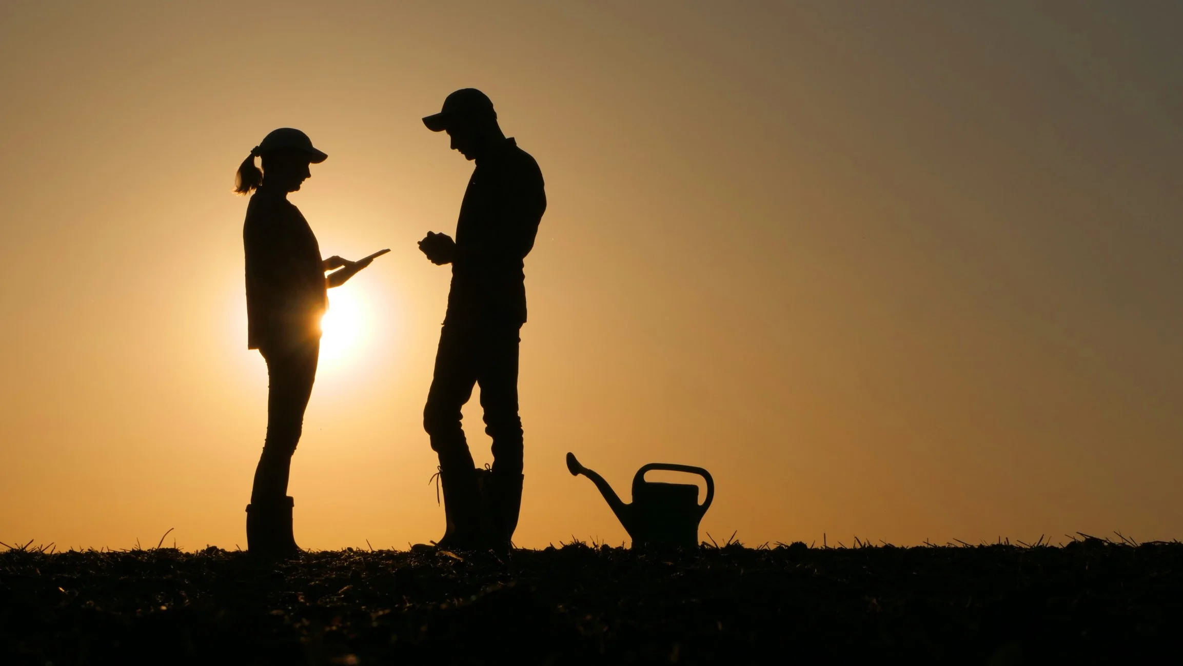 Silhouettes of two people, a man and a woman, standing outdoors during sunset, with a watering can nearby.
