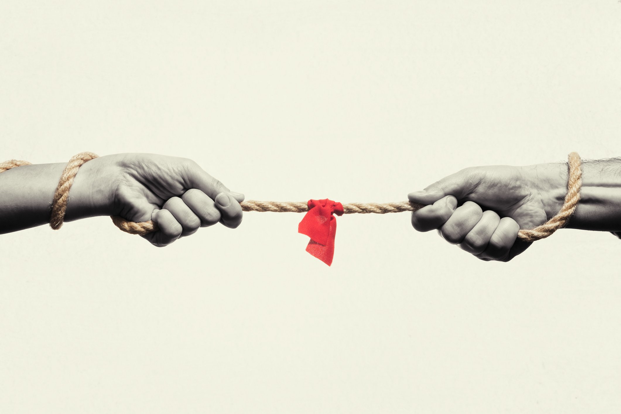 Two hands holding opposite ends of a rope with a red cloth tied in the middle, symbolizing conflict or division.