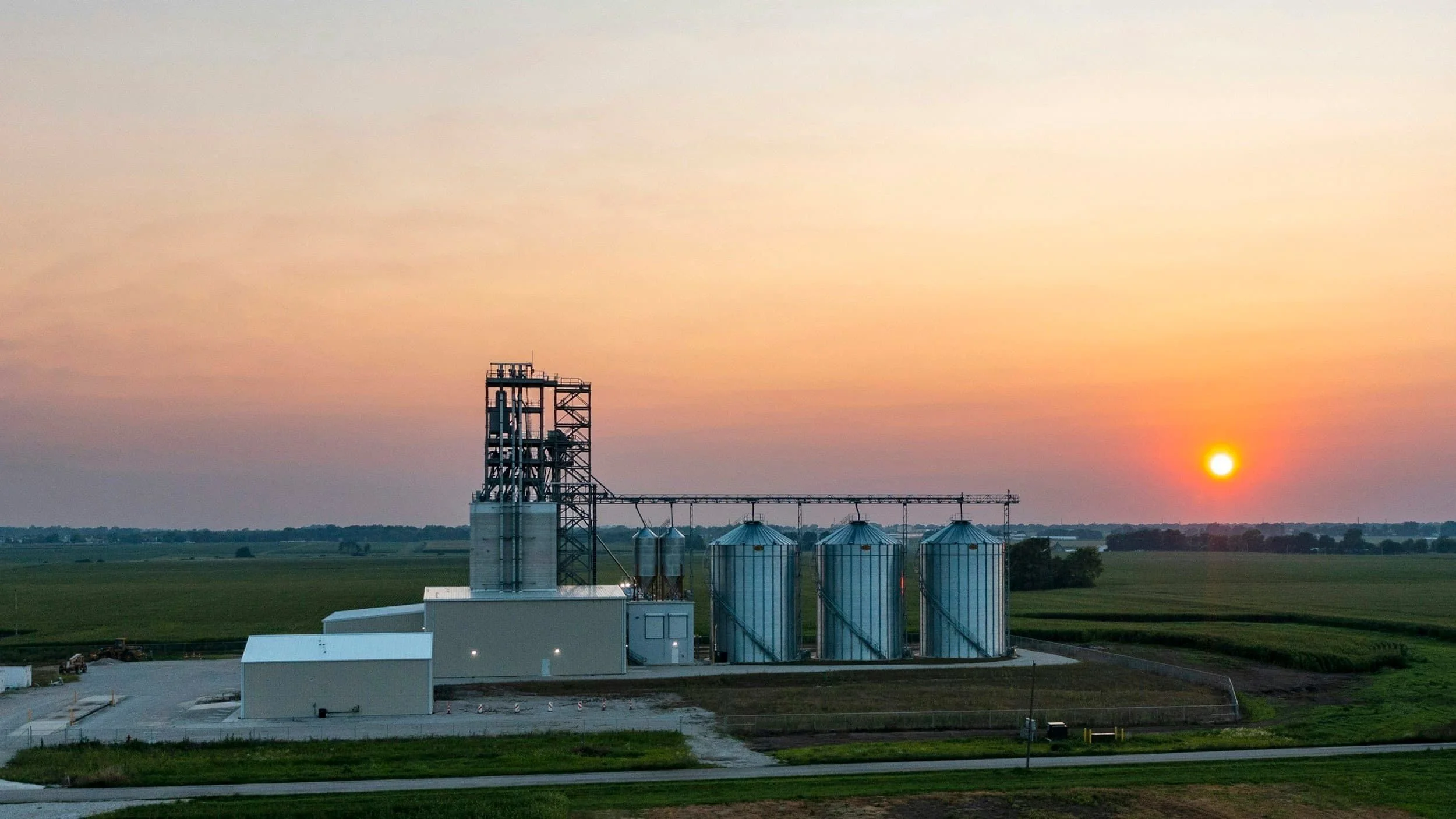A rural industrial facility with silos and metal structures during sunset with open fields in the background.