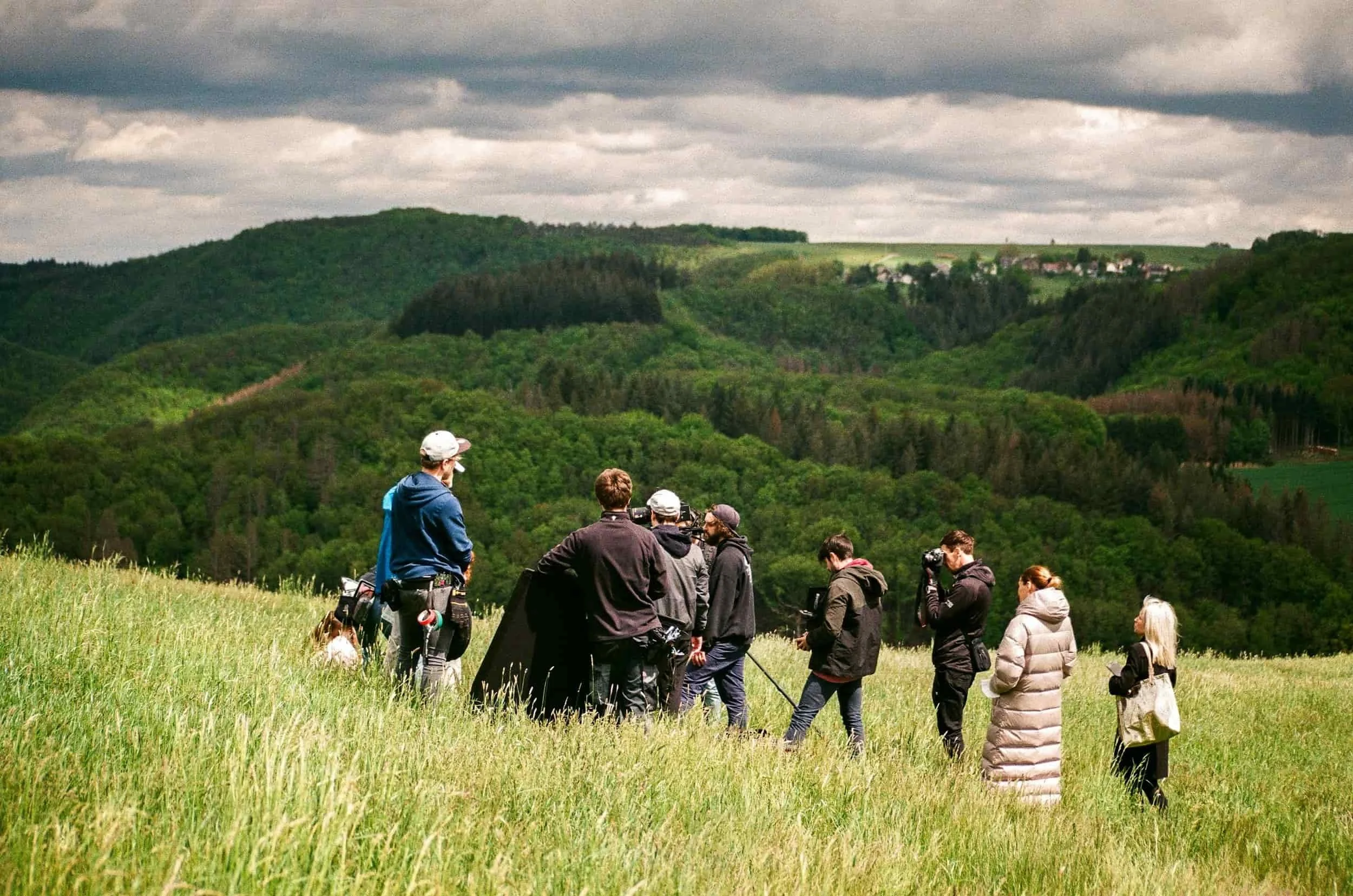 A group of people standing in a grassy field during cloudy weather, with lush green hills and a small village in the background.