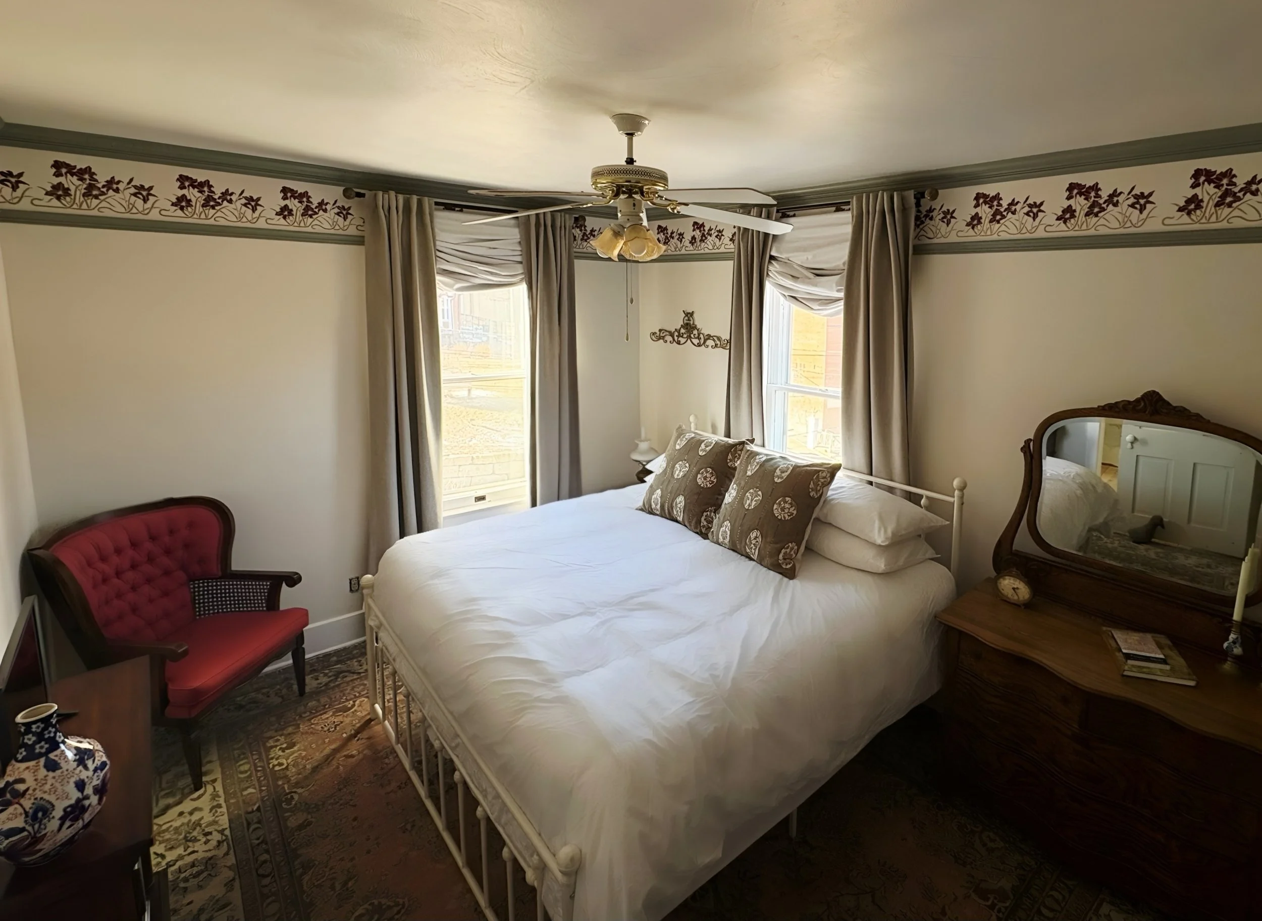 Peaceful and calming bedroom with King size bed, antique furniture, windows with draped curtains, a ceiling fan, and antique mirror. The room has a hand-painted floral border and antique floral patterned rug.