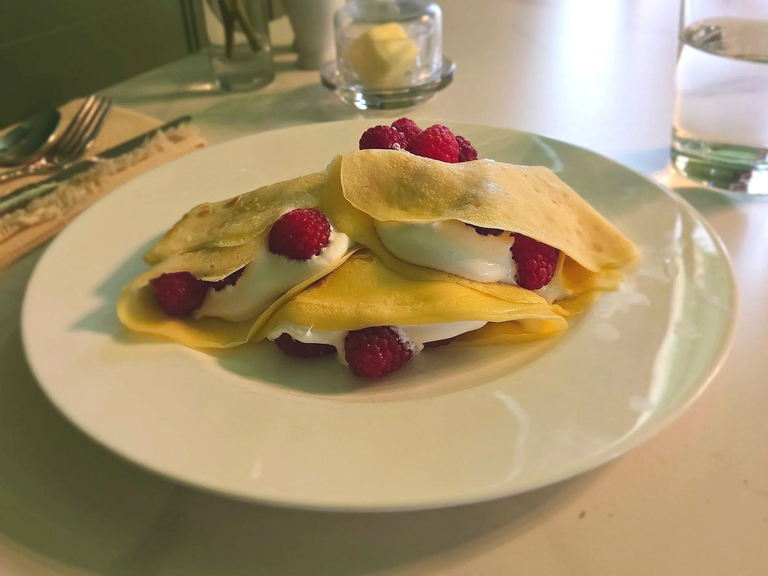 A delightful, light breakfast. Delicate crepes filled with creme and topped with raspberries, on a classic table set with a glassware, butter, and utensils.