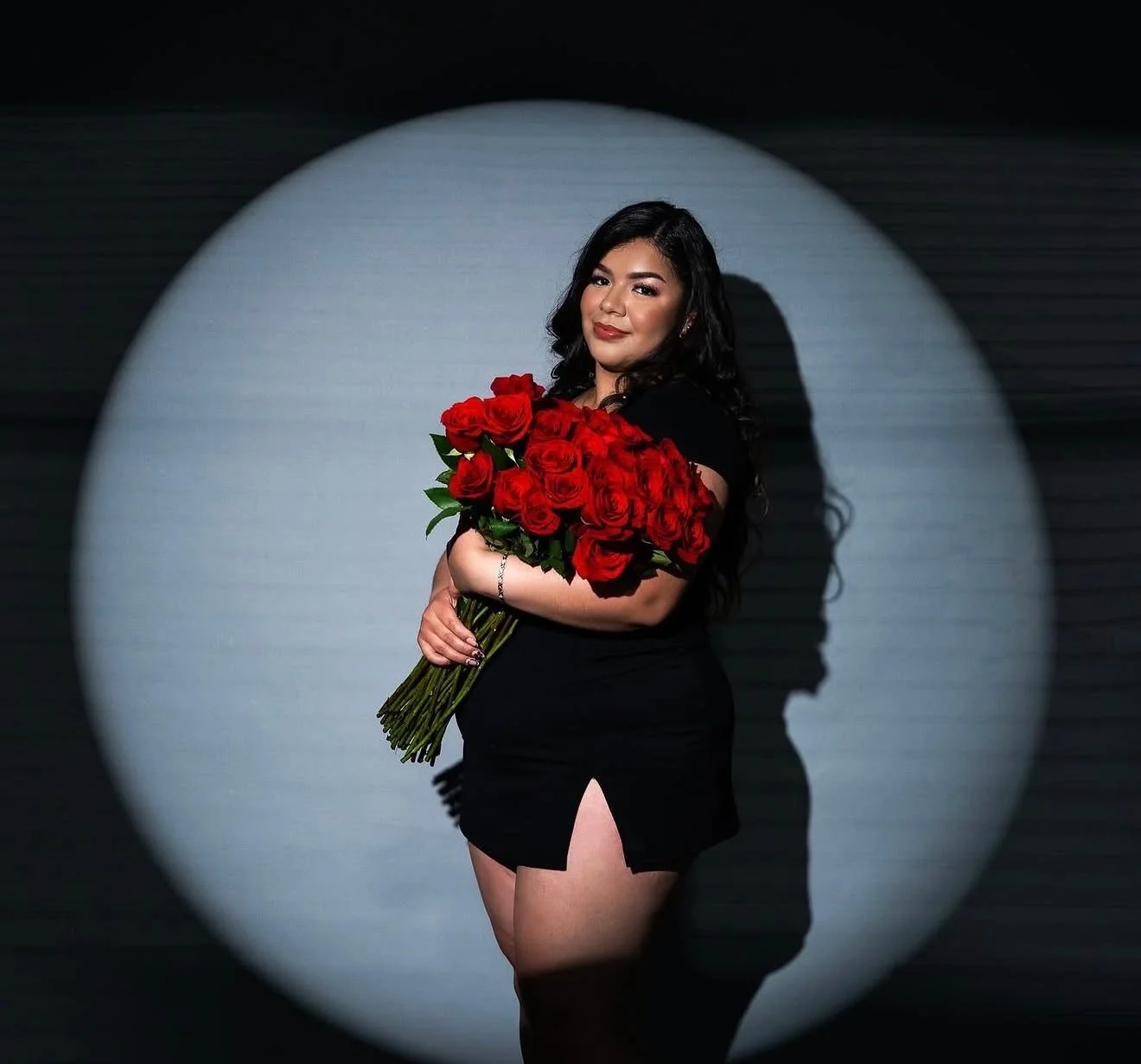 Woman in black dress holding a large bouquet of red roses, standing in front of a spotlight background.