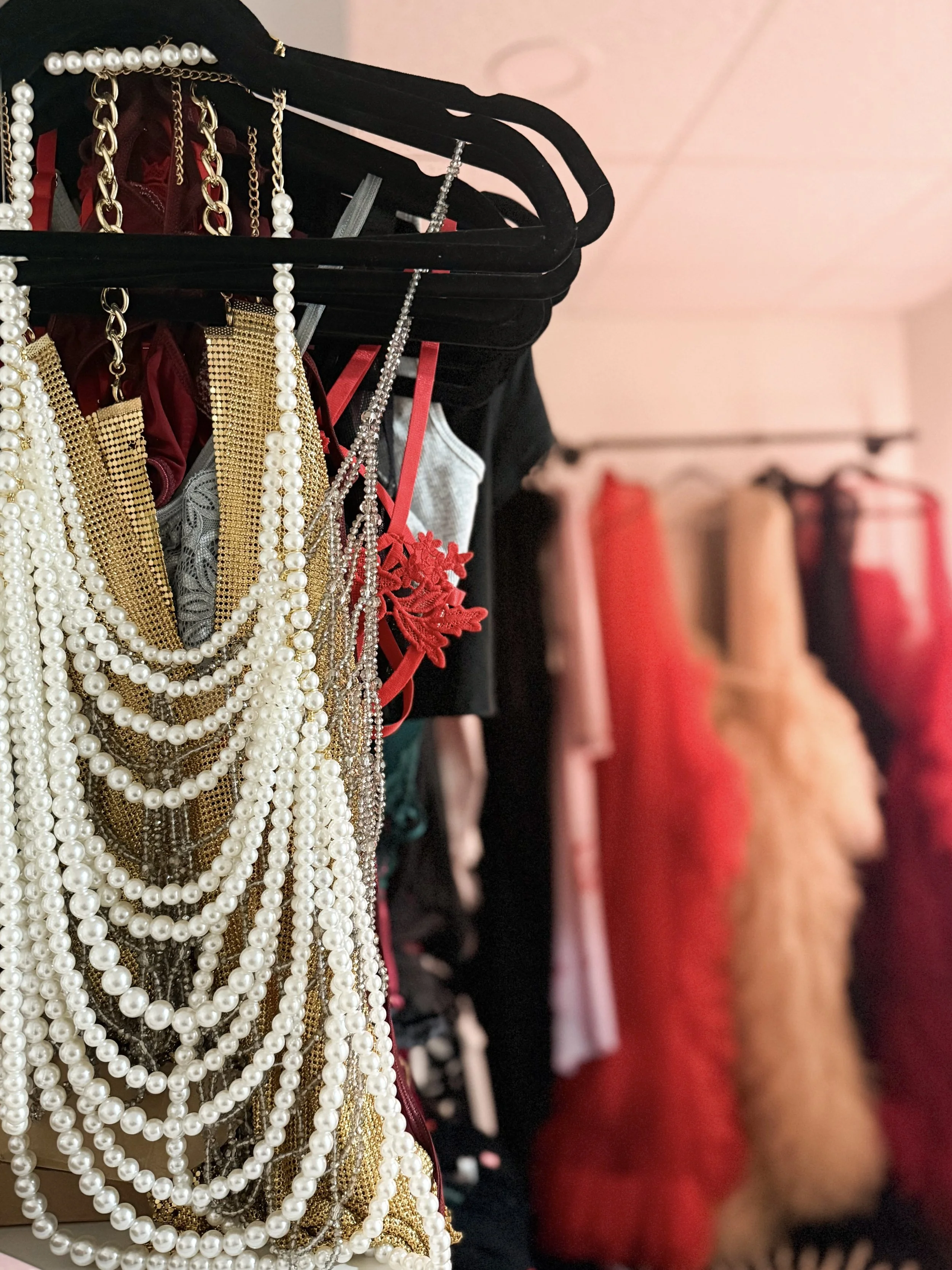 A display of jewelry, including pearl necklaces, gold chains, and silver chains, hanging on a black rack. In the background, various dresses and shoes are visible, some in red, pink, and beige colors.