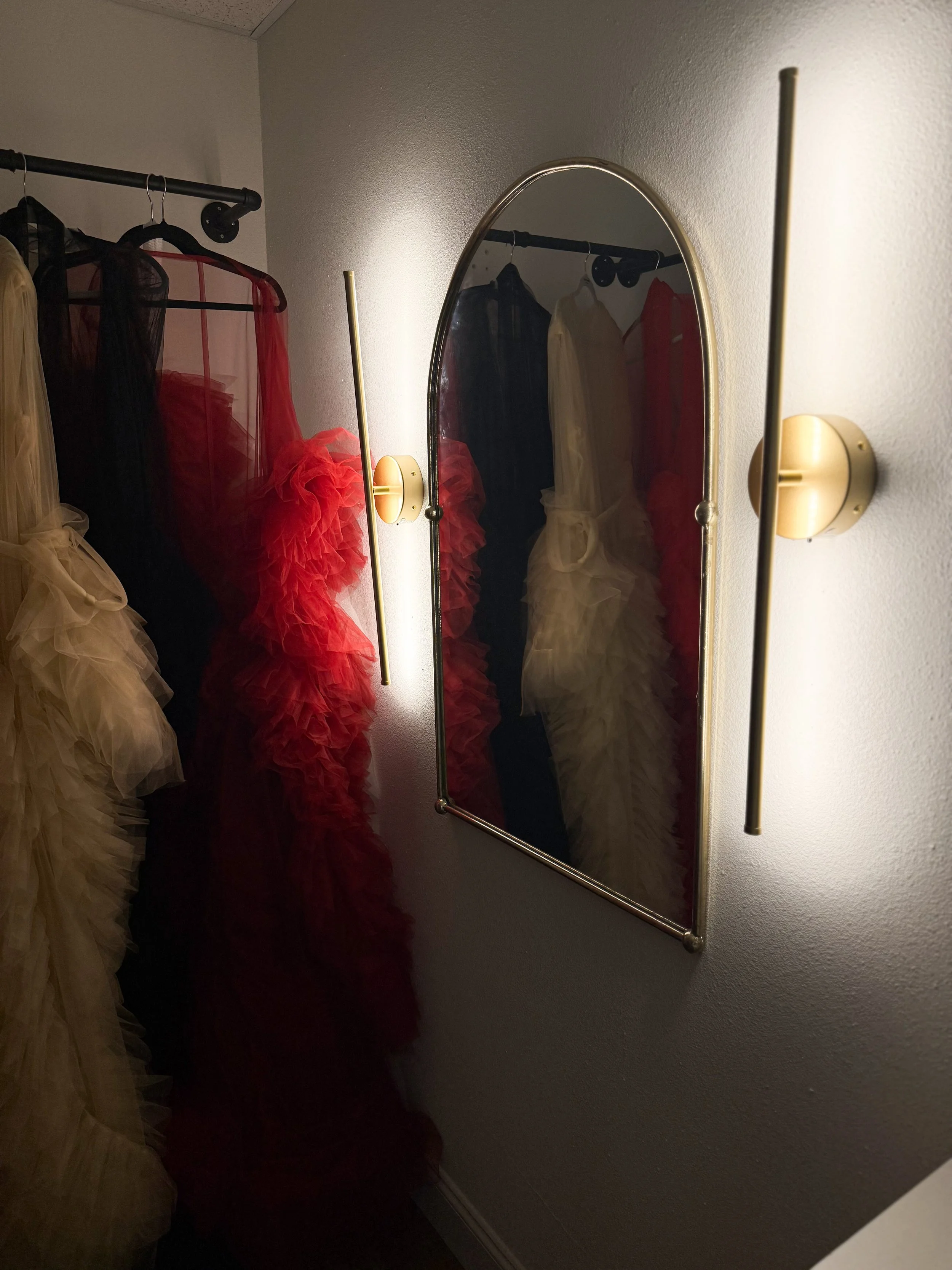 A closet with a mirror on the wall, showing dresses in various colors including beige, black, red, and cream, hanging on a black clothing rod. The room has a white textured wall and a light fixture with two gold-colored bars mounted on the wall beside the mirror.
