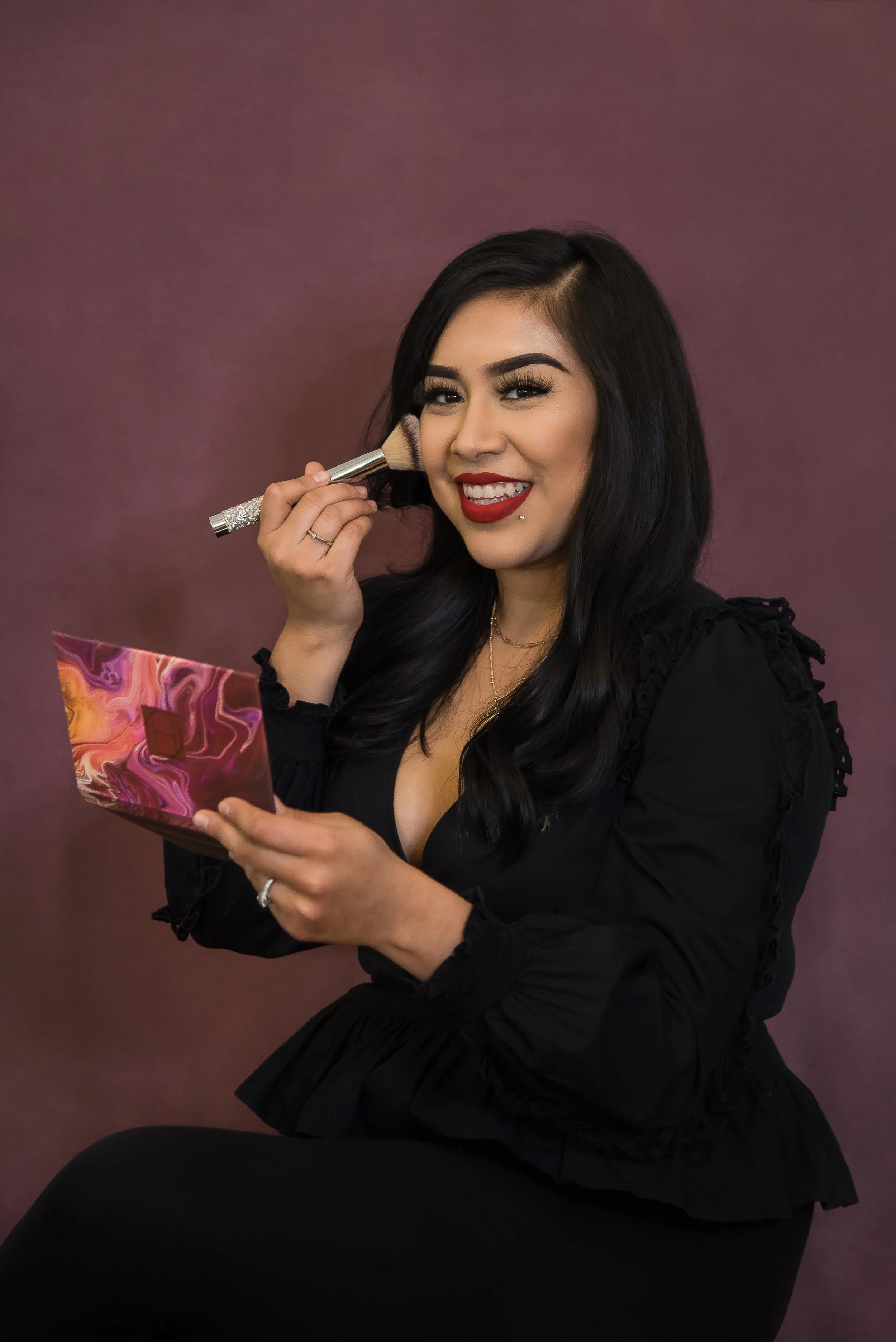 A woman with long black hair, wearing a black dress and red lipstick, sits on a black chair against a mauve background. She is smiling and applying makeup with a brush while holding a colorful makeup palette.