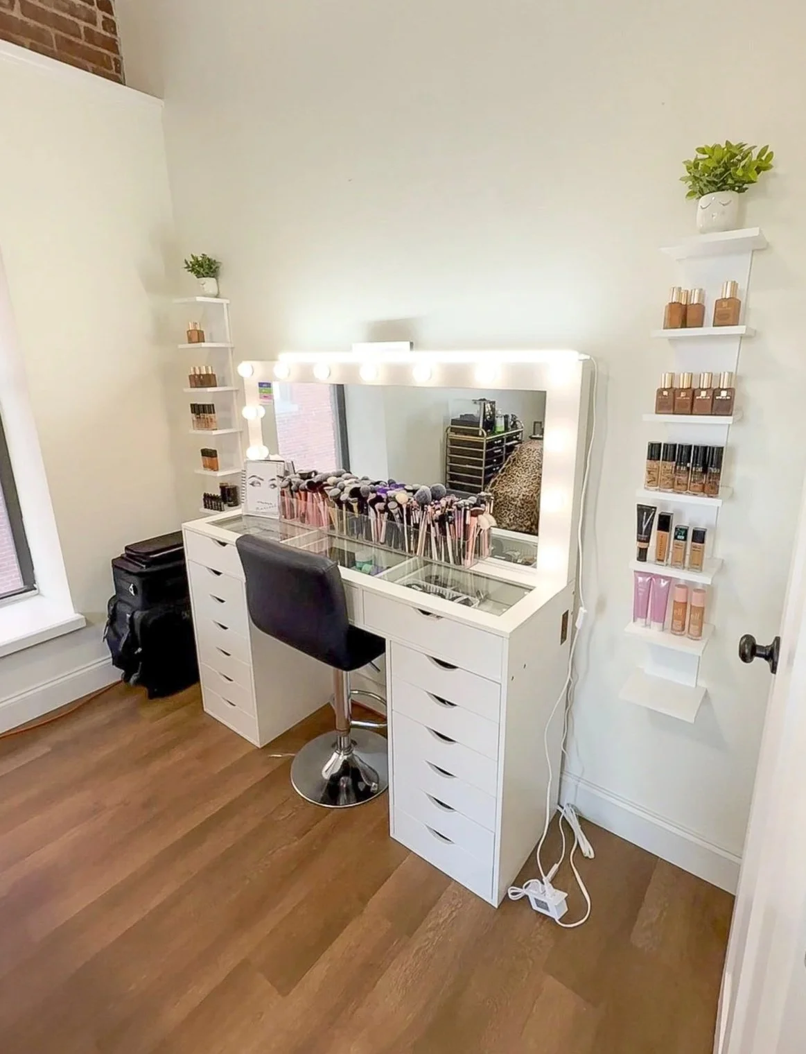 A makeup vanity setup with a large mirror surrounded by lights. The vanity has various makeup brushes and beauty products. Shelves on either side hold additional makeup and skincare items. A black chair sits in front of the vanity, and a window is visible to the left.
