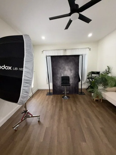 Photo of a photography studio setup with a black backdrop, a stool, a studio light, and some plants and furniture in a room with wooden floors and a ceiling fan.