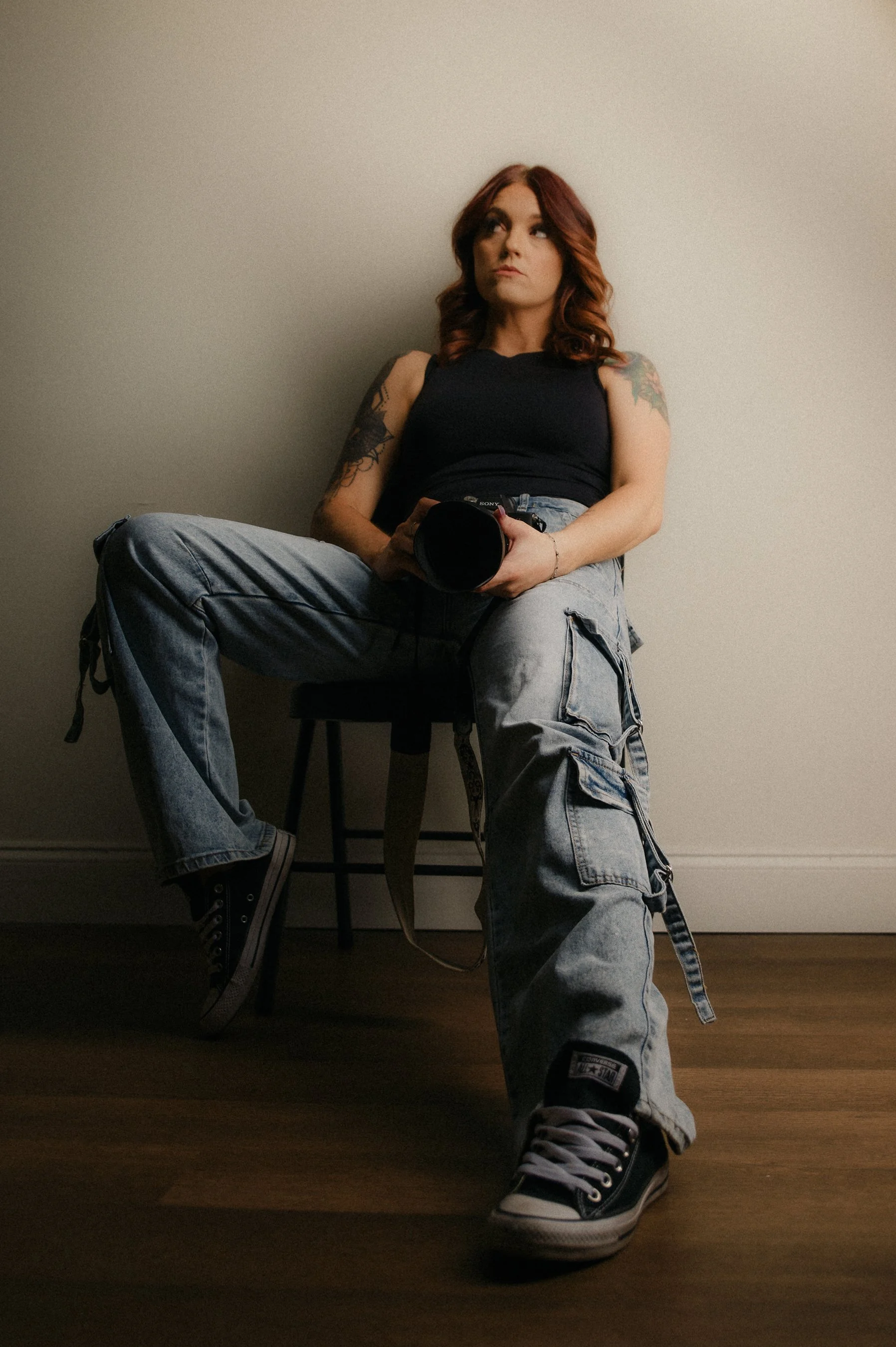 Young woman with red hair, tattoos, black tank top, baggy jeans, and black sneakers seated against a plain wall, holding a camera.