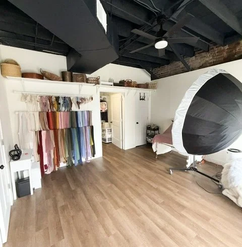 Photo of a photo studio with fabric swatches hanging on a wall, a large umbrella light, and a bed with pink bedding.