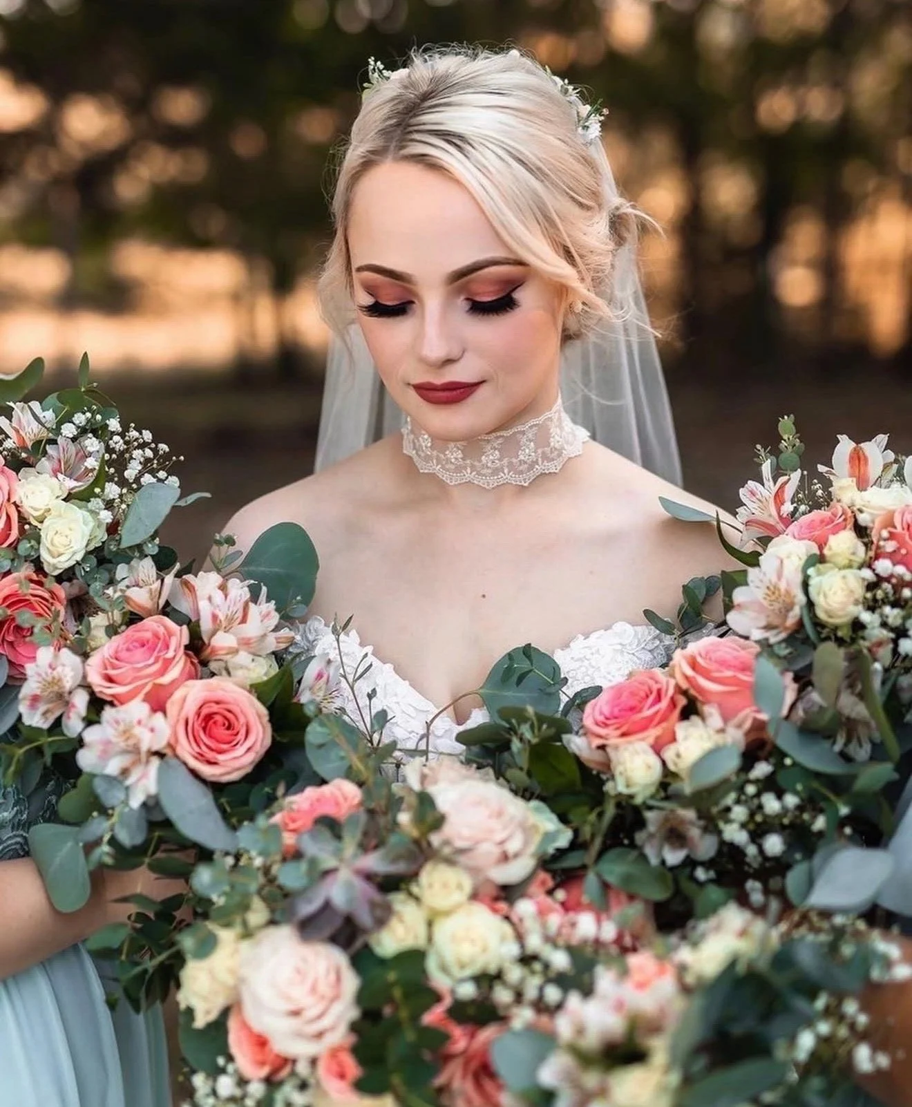 A young woman in wedding attire, with blonde hair and makeup, is standing outdoors among a large bouquet of pink and white roses and greenery, with trees and a sunset in the background.