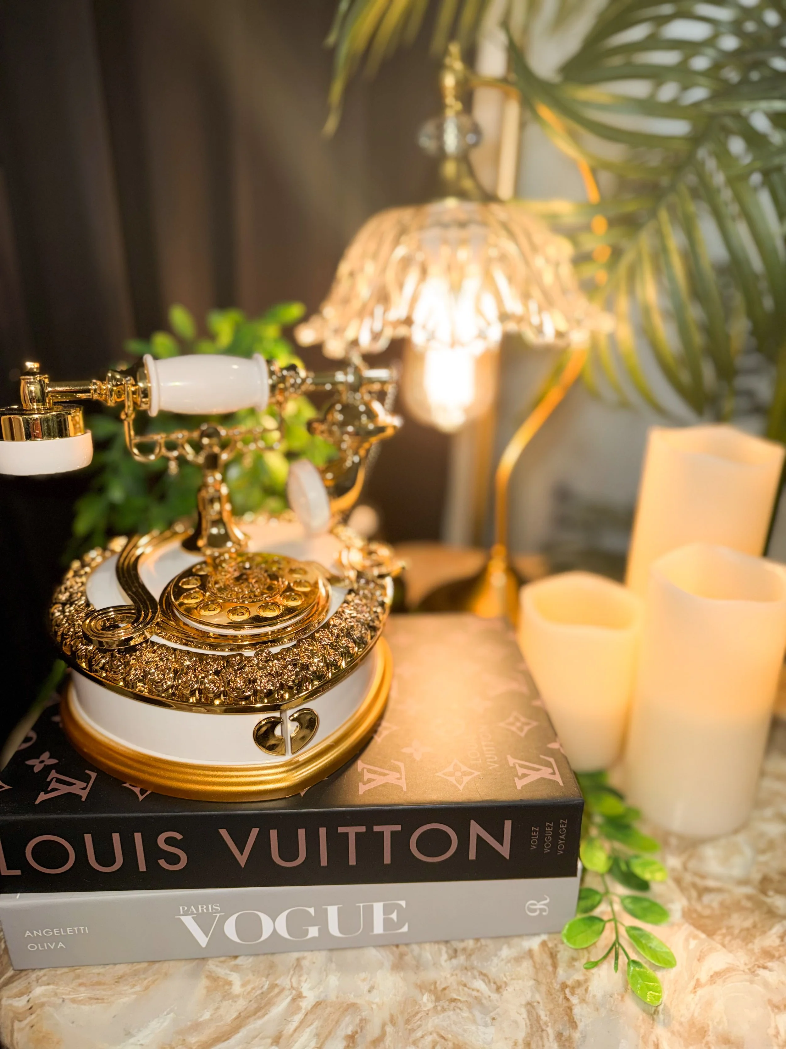 A vintage gold and white jewelry box with a decorative lid, possibly containing jewelry, placed on top of fashion magazine books titled "VOGUE". In the background, there is a lit lamp with a shade, green leafy plants, and artificial candles on a marble surface.