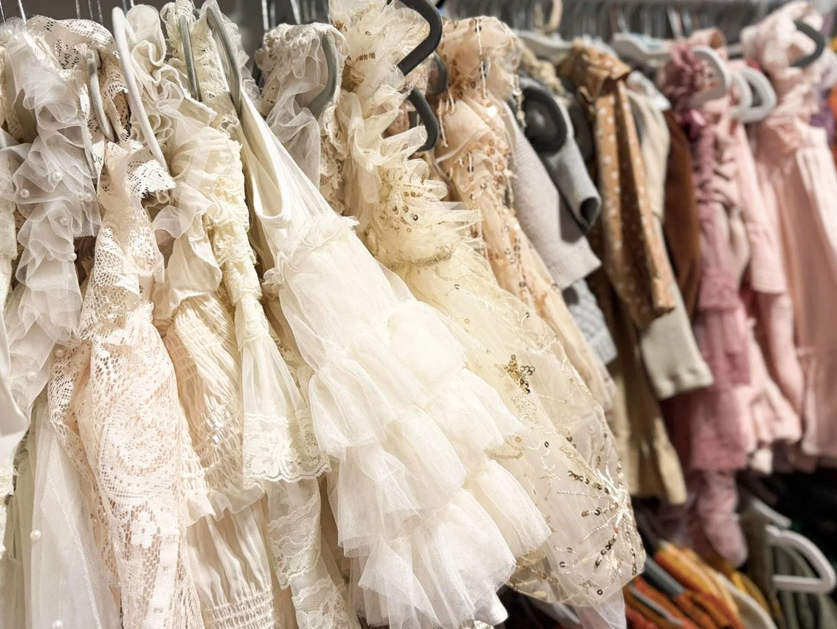 A row of children's dresses in varying shades of white and pink hanging on a rack, featuring lace, tulle, and decorative embellishments.
