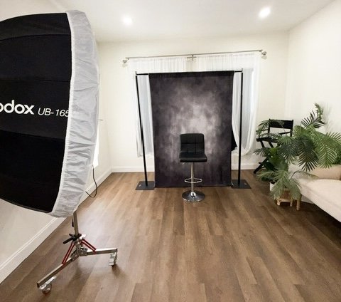Photography studio setup with a black backdrop, professional lighting softbox on a stand, and a black stool on a wooden floor, with a couch and plants in the background.