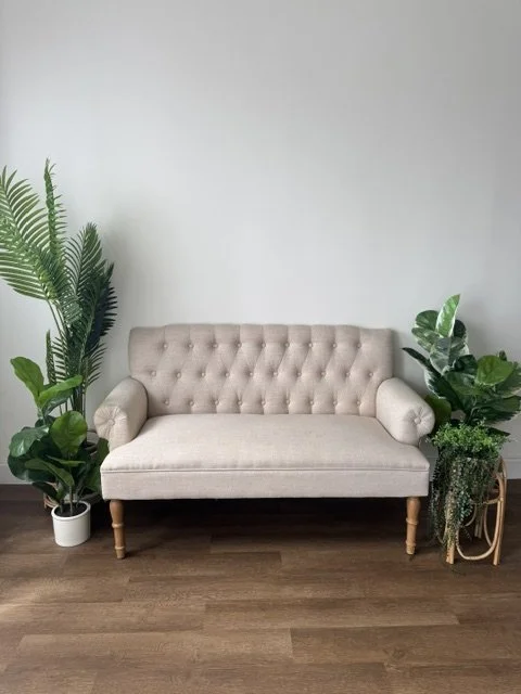 A beige tufted sofa with rolled arms in a room with a wooden floor, flanked by large green potted plants on either side.