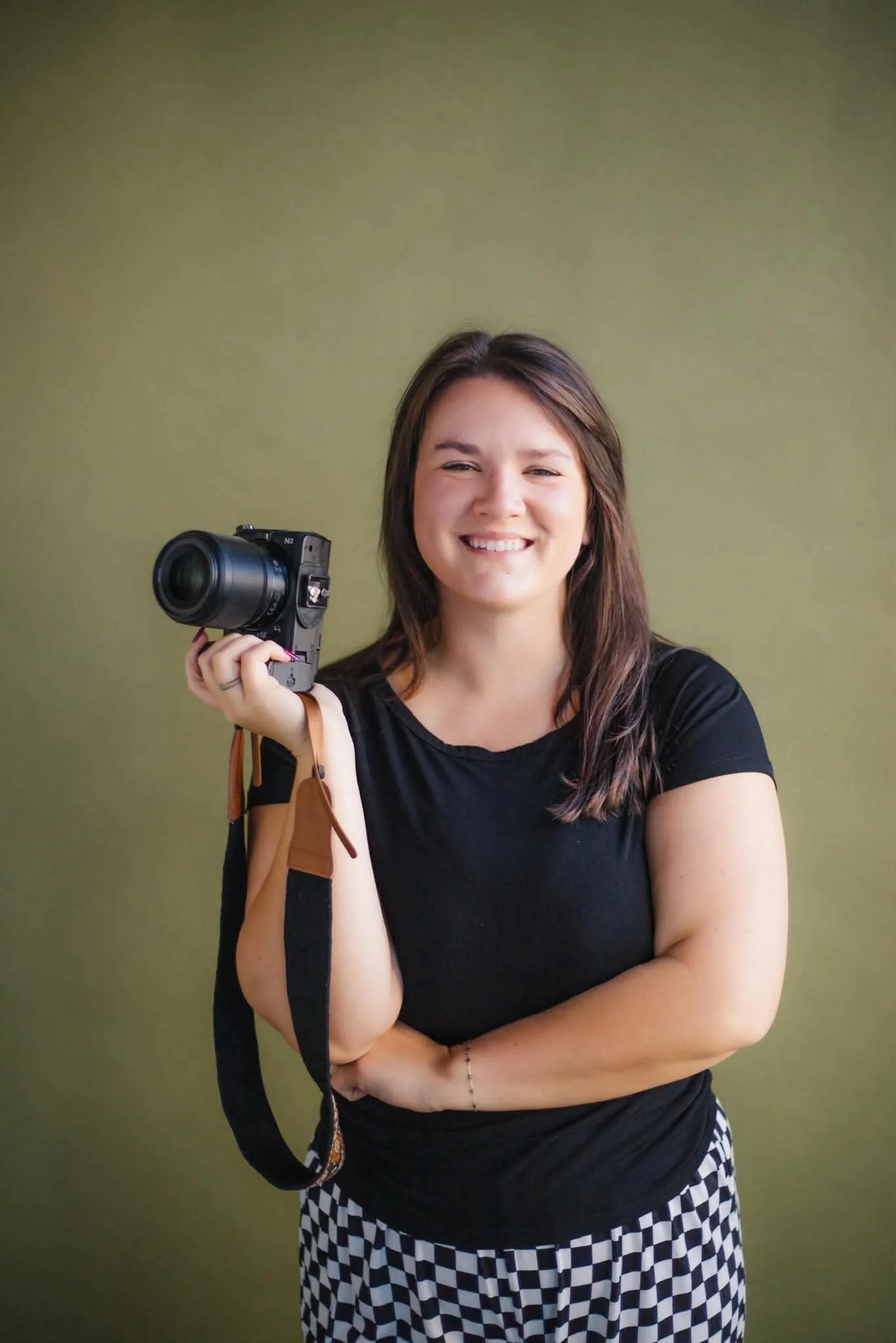 A young woman with long brown hair smiling and holding a camera in her right hand. She is wearing a black t-shirt and checkered black and white pants, standing against a plain green background.