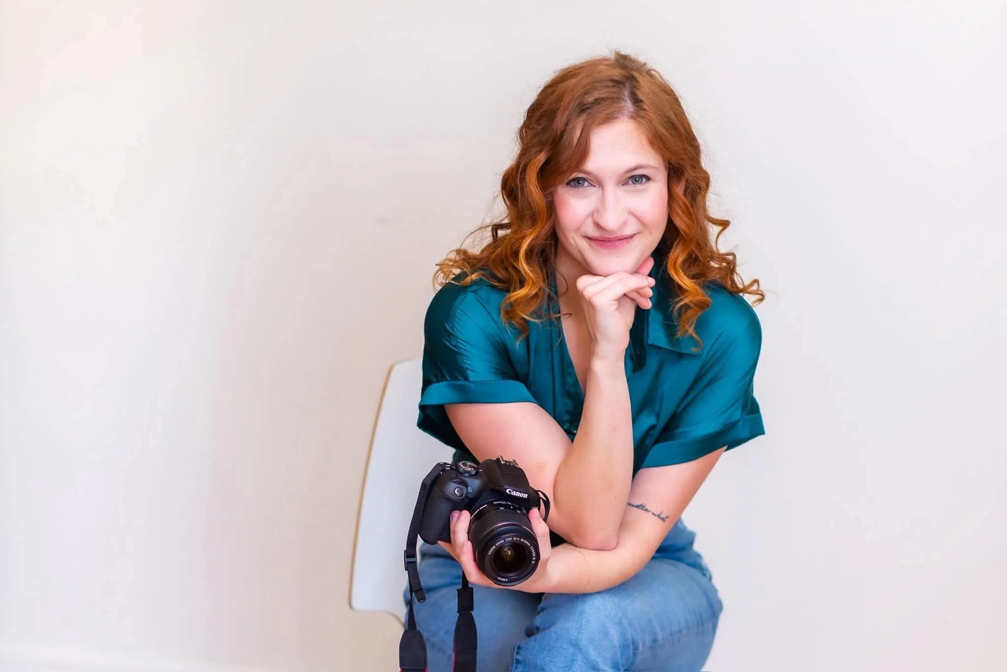 A woman with red hair and blue eyes sitting with a camera in her hands against a plain white background, smiling at the camera.