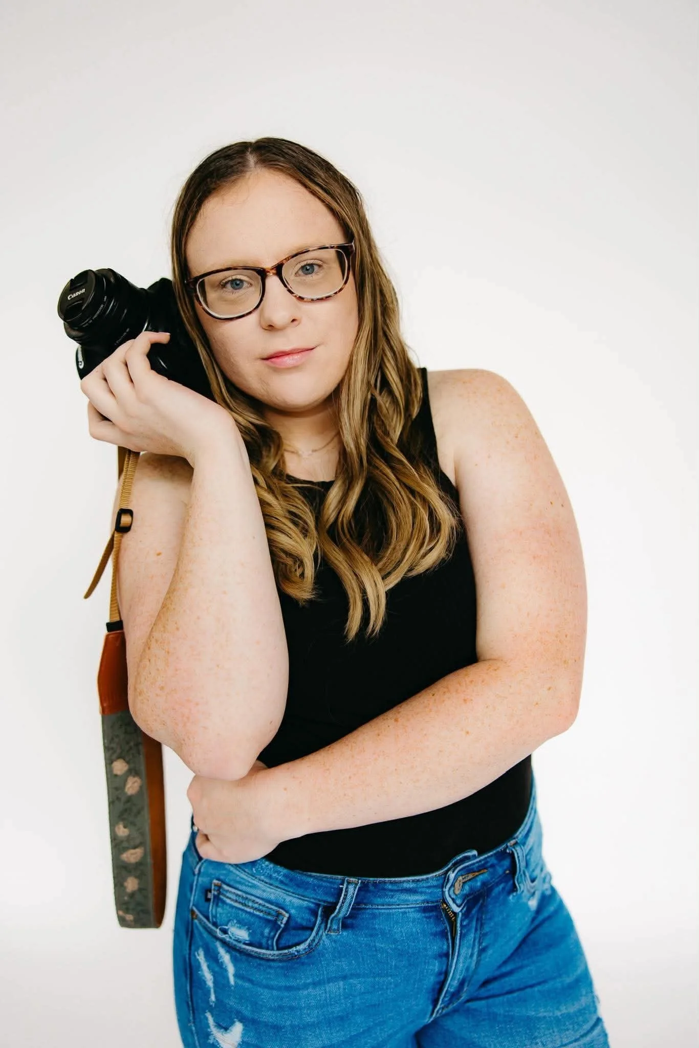 A woman with long wavy hair, glasses, and light skin posing against a plain white background. She's wearing a black sleeveless top, blue jeans, and carrying a patterned shoulder bag. She is holding a Canon camera to her ear, giving a confident expression.