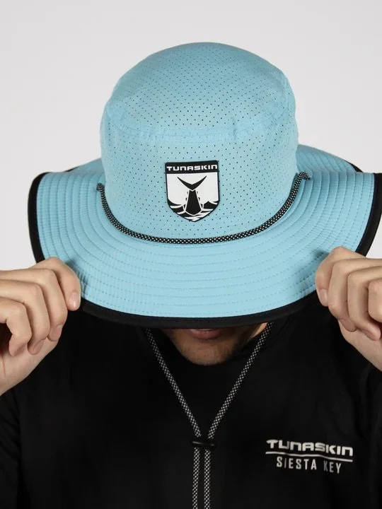 Person wearing a light blue wide-brimmed hat with the Tunaskin logo, holding the brim with both hands, black shirt with Tunaskin Siesta Key logo.