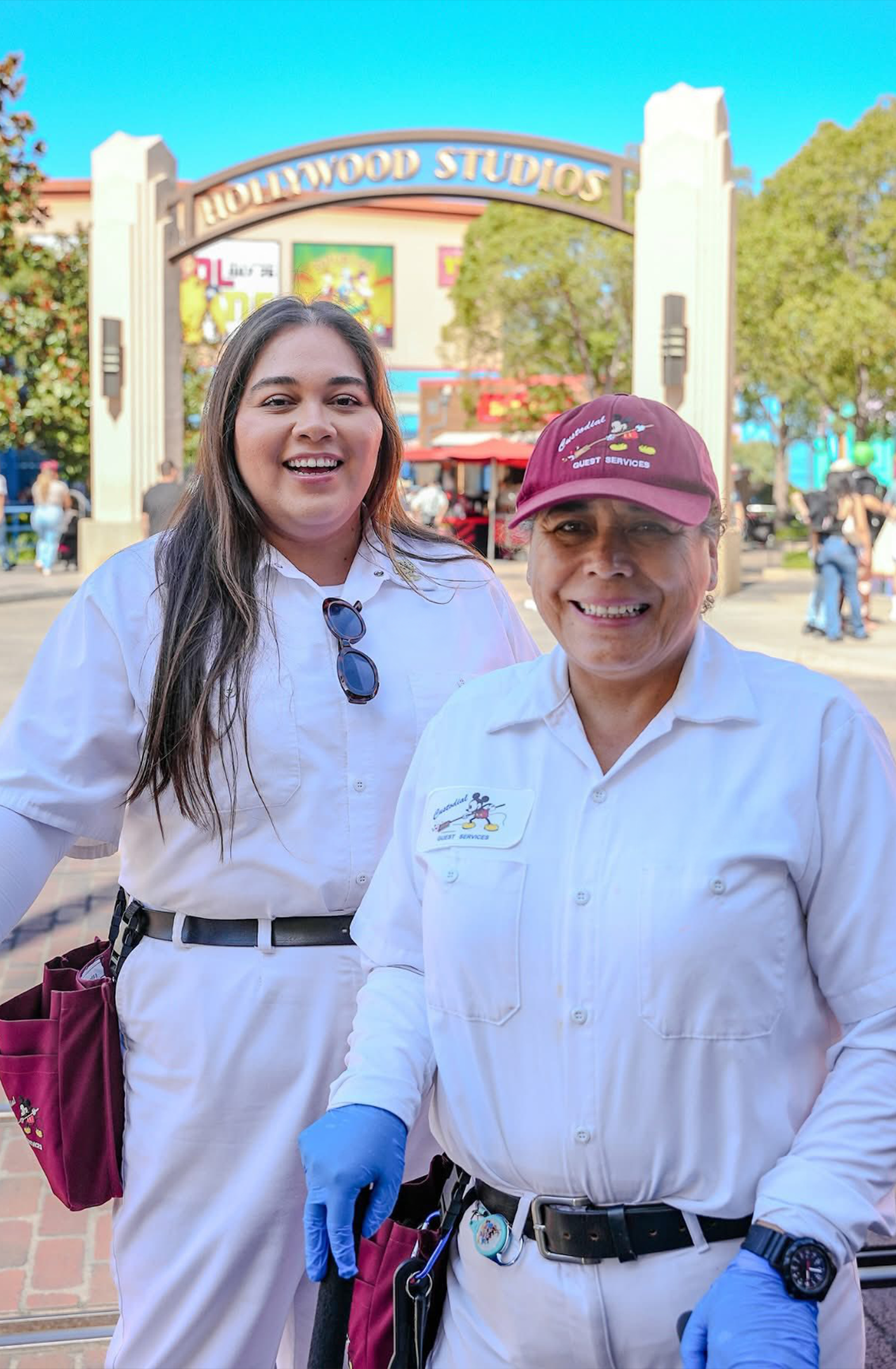 Two women in white uniforms standing outdoors in front of Hollywood Studios entrance sign. One woman is wearing a maroon cap, and both are smiling, with the woman on the left holding her hand out, wearing a glove, possibly preparing for a health-rela