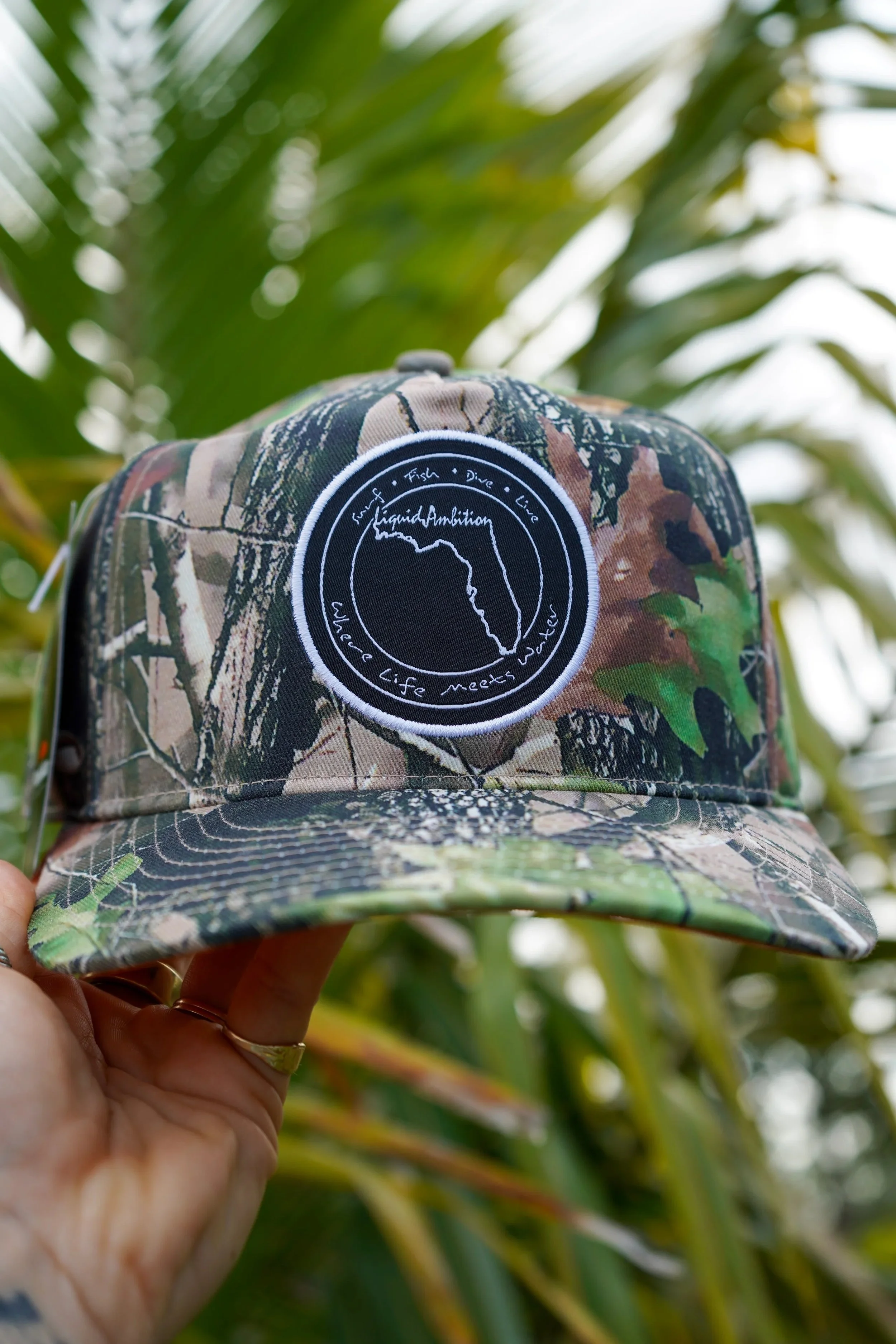 A camouflage baseball cap with a circular patch on the front showing an outline of the state of Florida. The patch has the words "Liquid Ambition" and "where life meets water". The background is blurry green foliage.