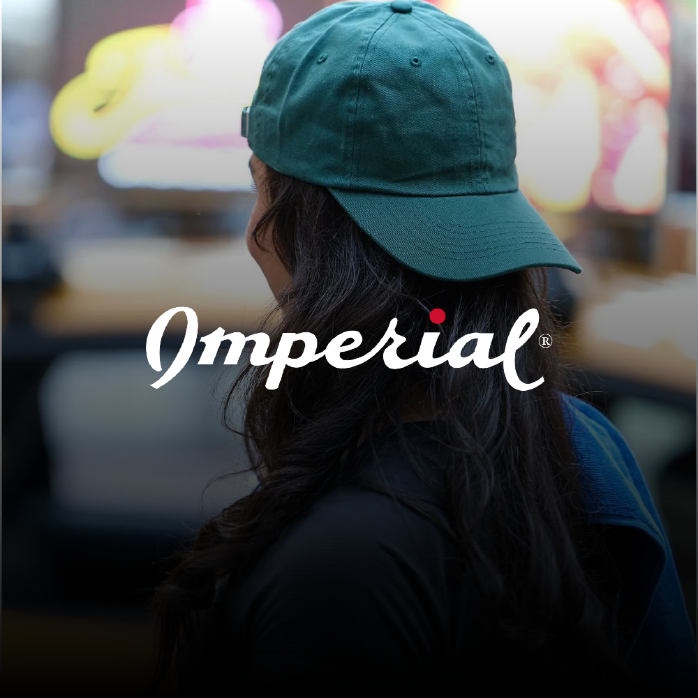 A woman wearing a teal baseball cap with her dark hair flowing over her shoulders, in a setting with blurred colorful lights in the background, featuring the word 'Imperial' in white cursive font with a red dot over the letter 'i'.
