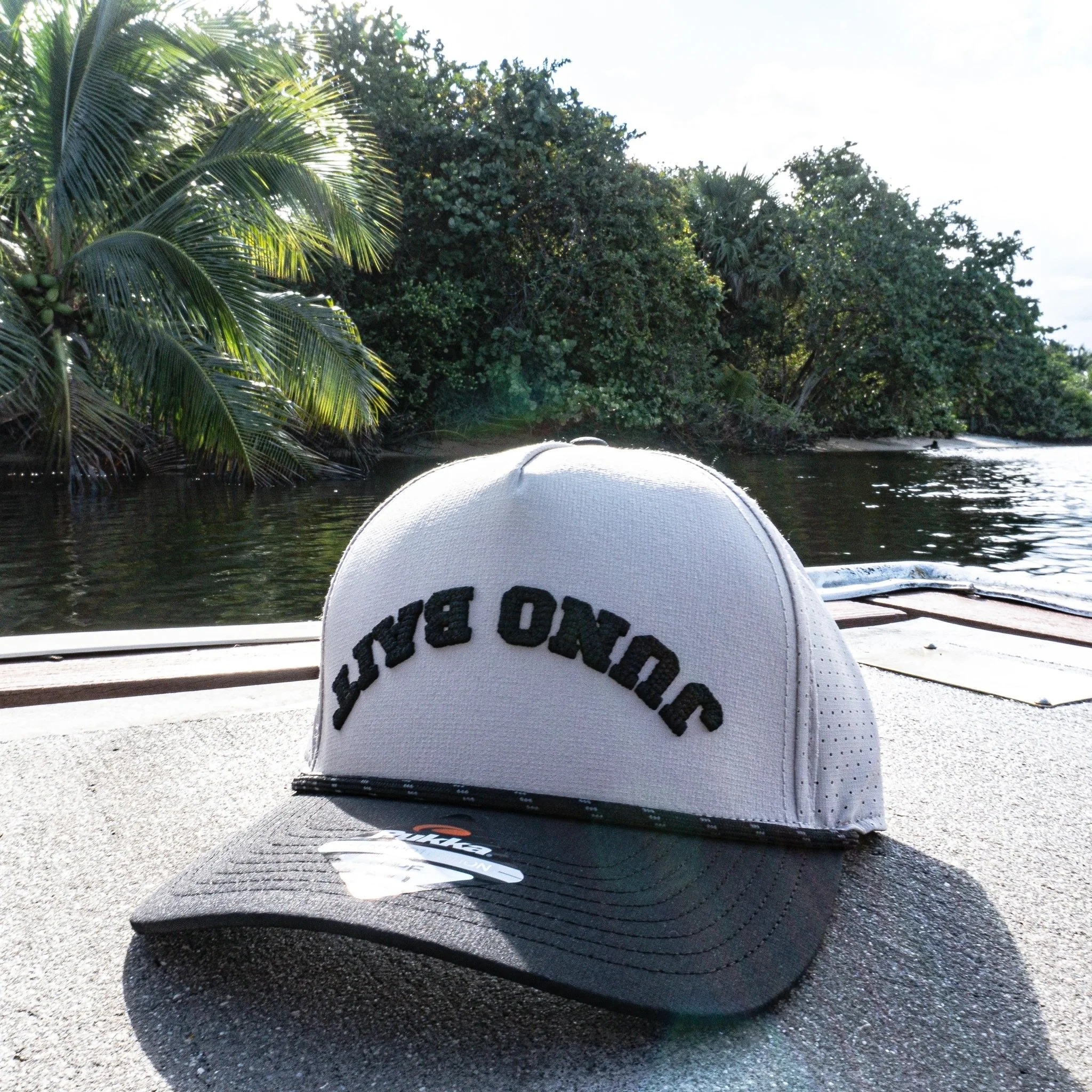 View from a boat on a river or lake, with a person's white hat in the foreground, green tropical trees and bushes along the shoreline, and a partly cloudy sky overhead.