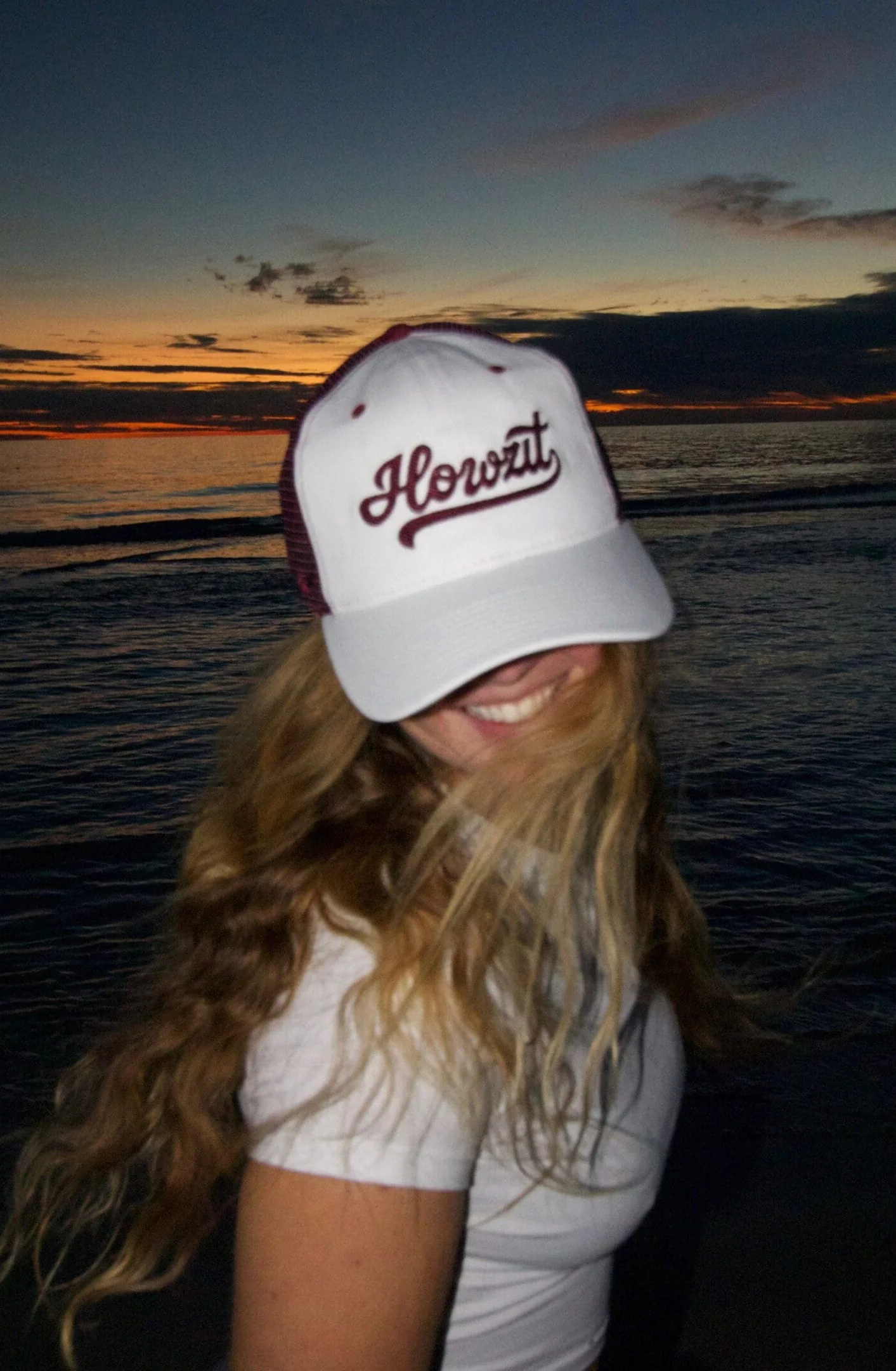 A woman with long, curly hair wearing a white cap with the word "Howzit" in red cursive, smiling at sunset by the water.