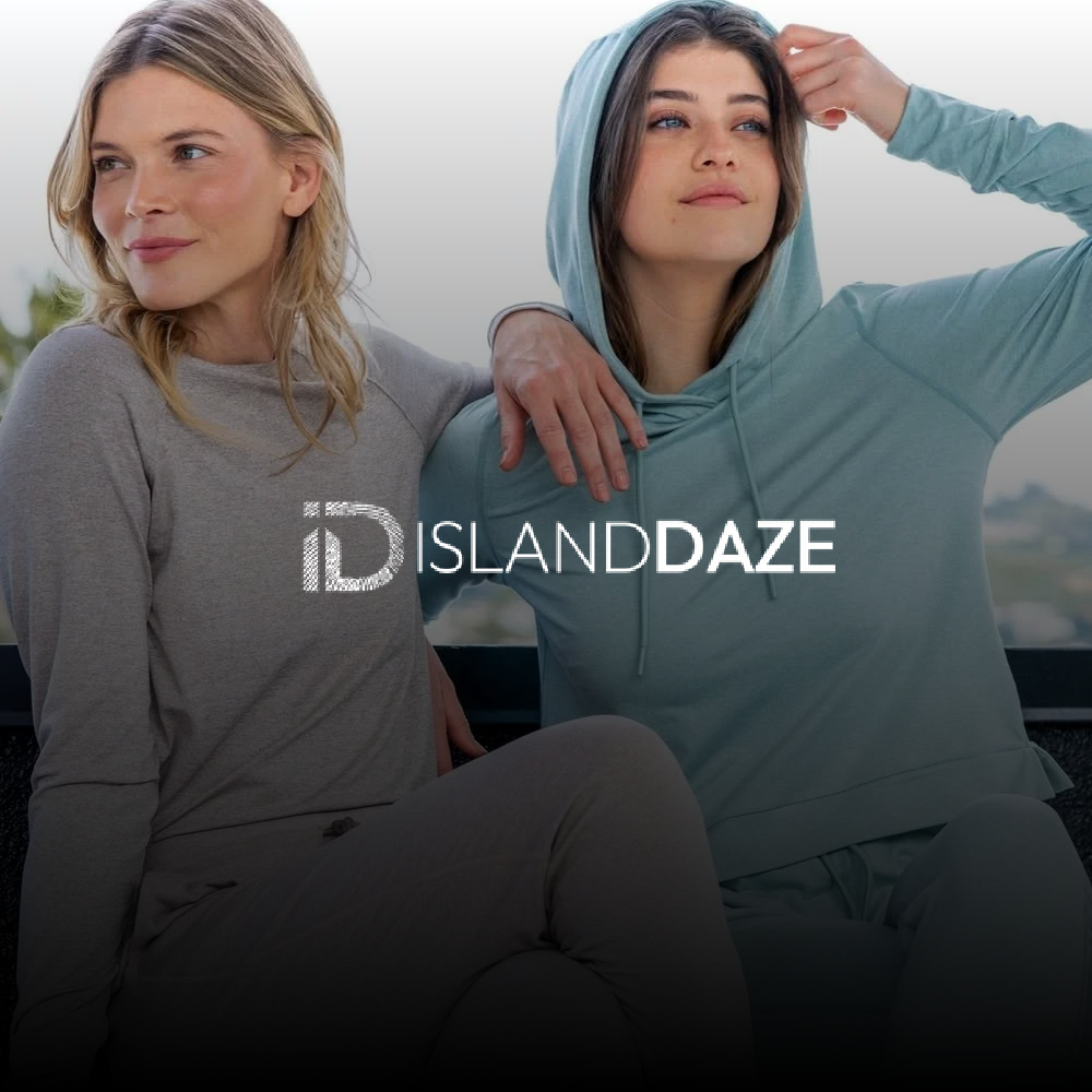 Two women sitting outdoors, one with blonde hair wearing a gray top, and the other with brown hair wearing a light blue hoodie. The woman in the hoodie is touching her hair and looking into the distance. The background is blurry with a hint of green trees and sky.