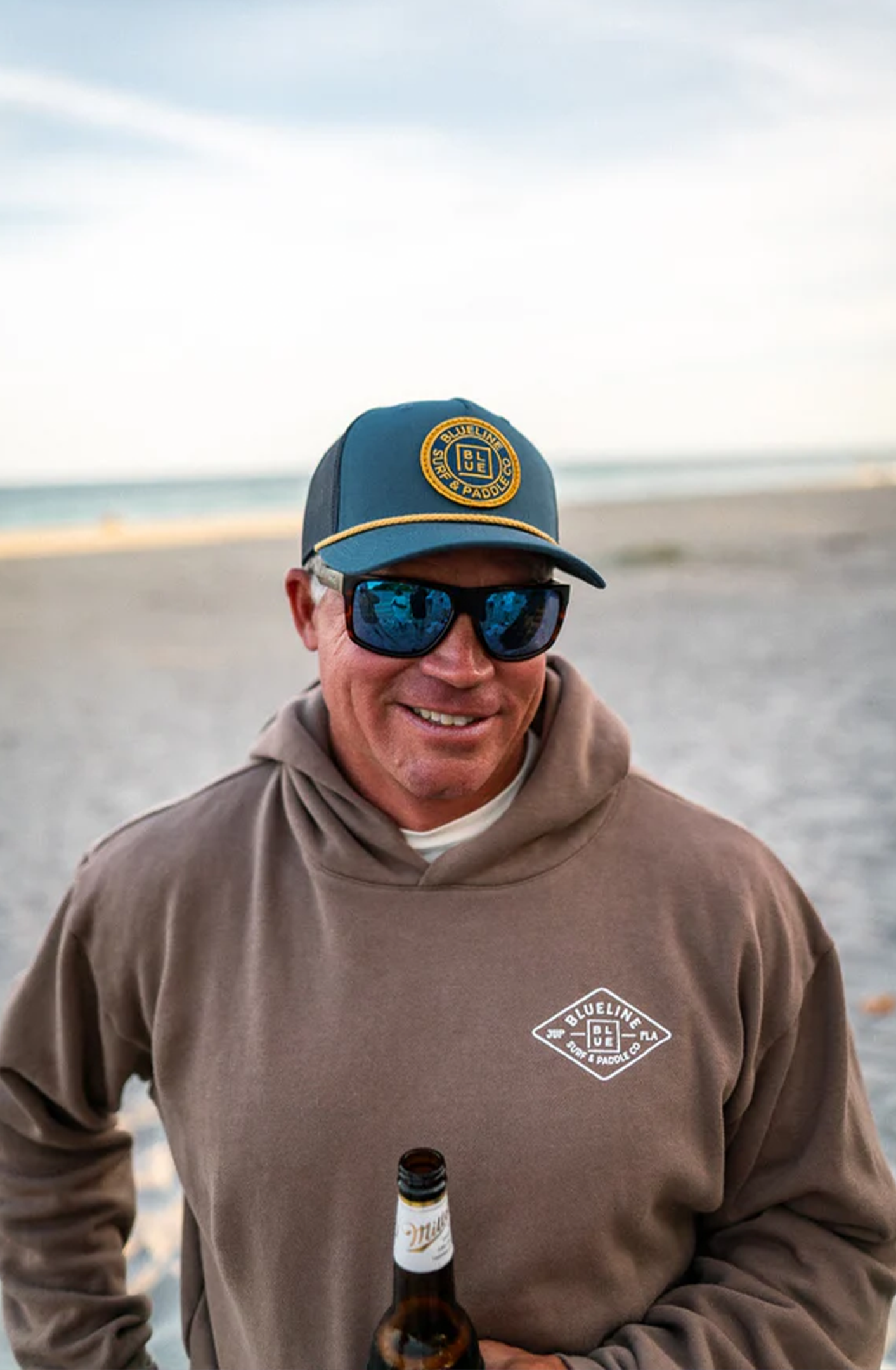 A man smiling on a beach, wearing sunglasses, a blue cap with a logo, and a brown hoodie with a logo, holding a beer bottle.