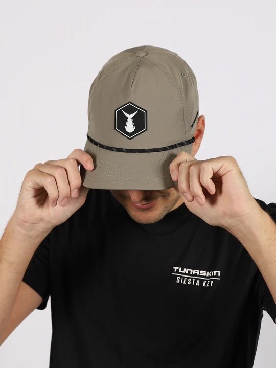 A man wearing a black T-shirt and a beige cap with a patch logo is adjusting the cap, with the logo depicting a fish in a pentagon shape.