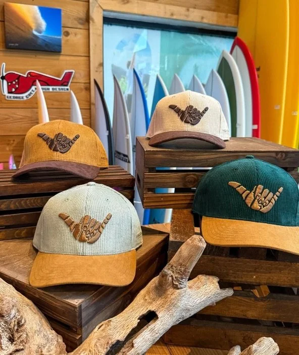 Four baseball caps with a shaka hand gesture embroidered on the front, displayed on wooden stands in a surf shop, with surfboards and wooden wall decor in the background.
