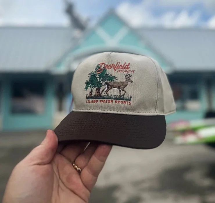 A beige and black cap with embroidered palm trees and a deer, with the text 'Pinefield Beach' and 'Island Water Sports' in front of an out-of-focus building and kayaks in the background.