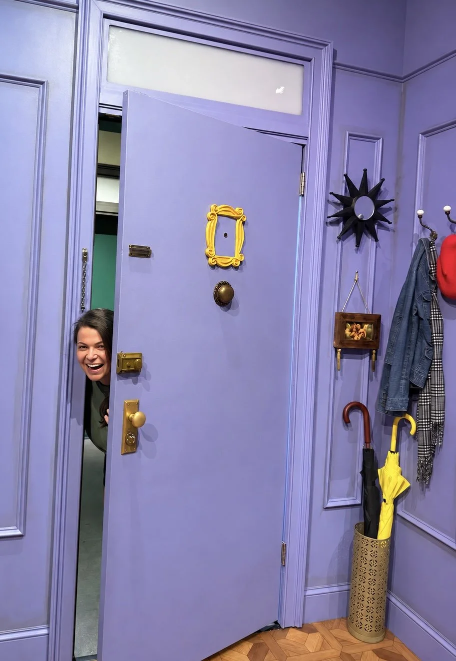 A woman peeking out of a partially open purple door with a cheerful expression. The room has purple walls with decorative molding, and there are umbrellas and coats hanging near the door.