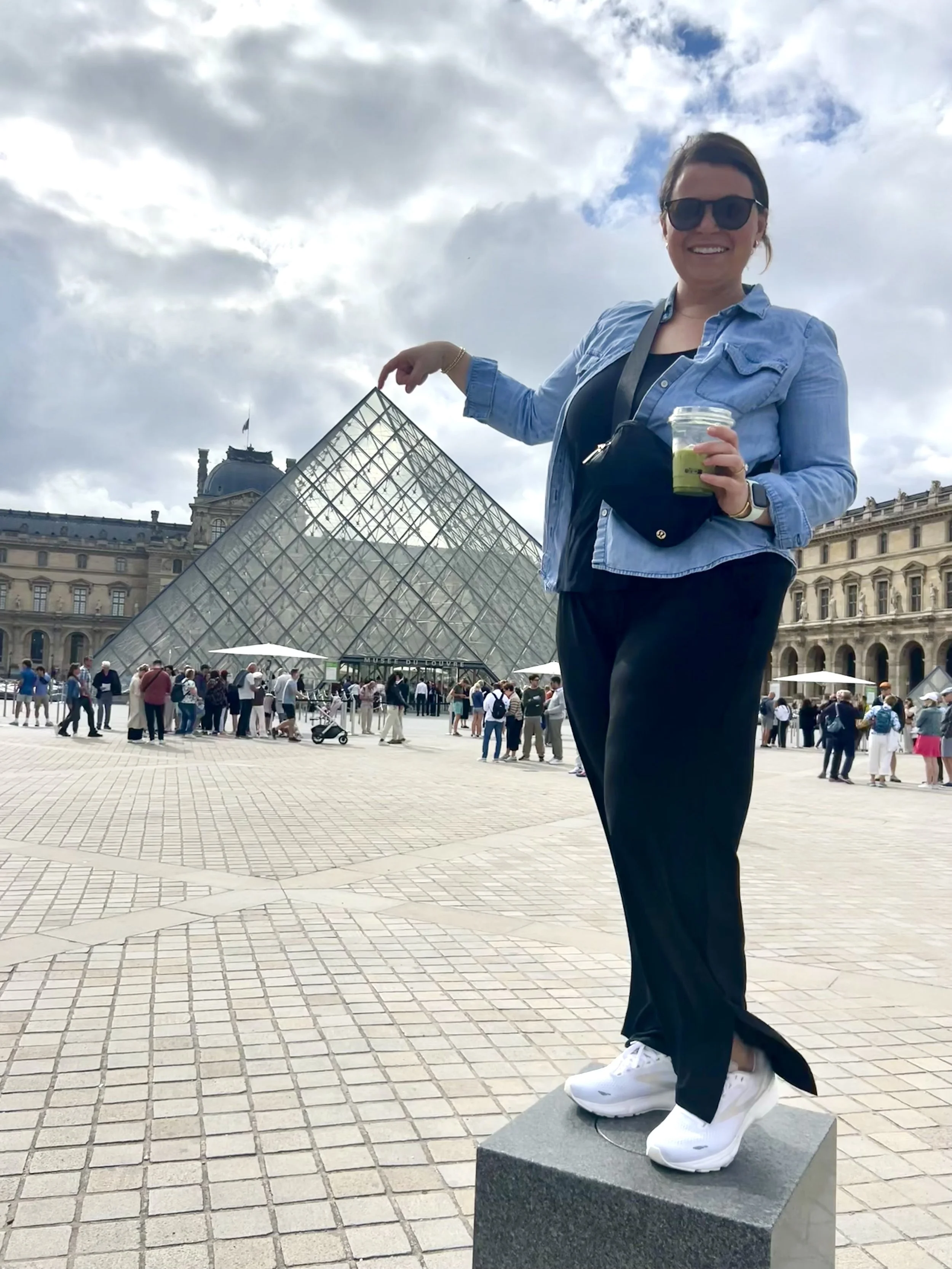 Woman smiling and pointing at the Louvre Pyramid in Paris, France, while standing on a stone pedestal, holding a drink, with a cloudy sky above and a crowd of tourists in the background.
