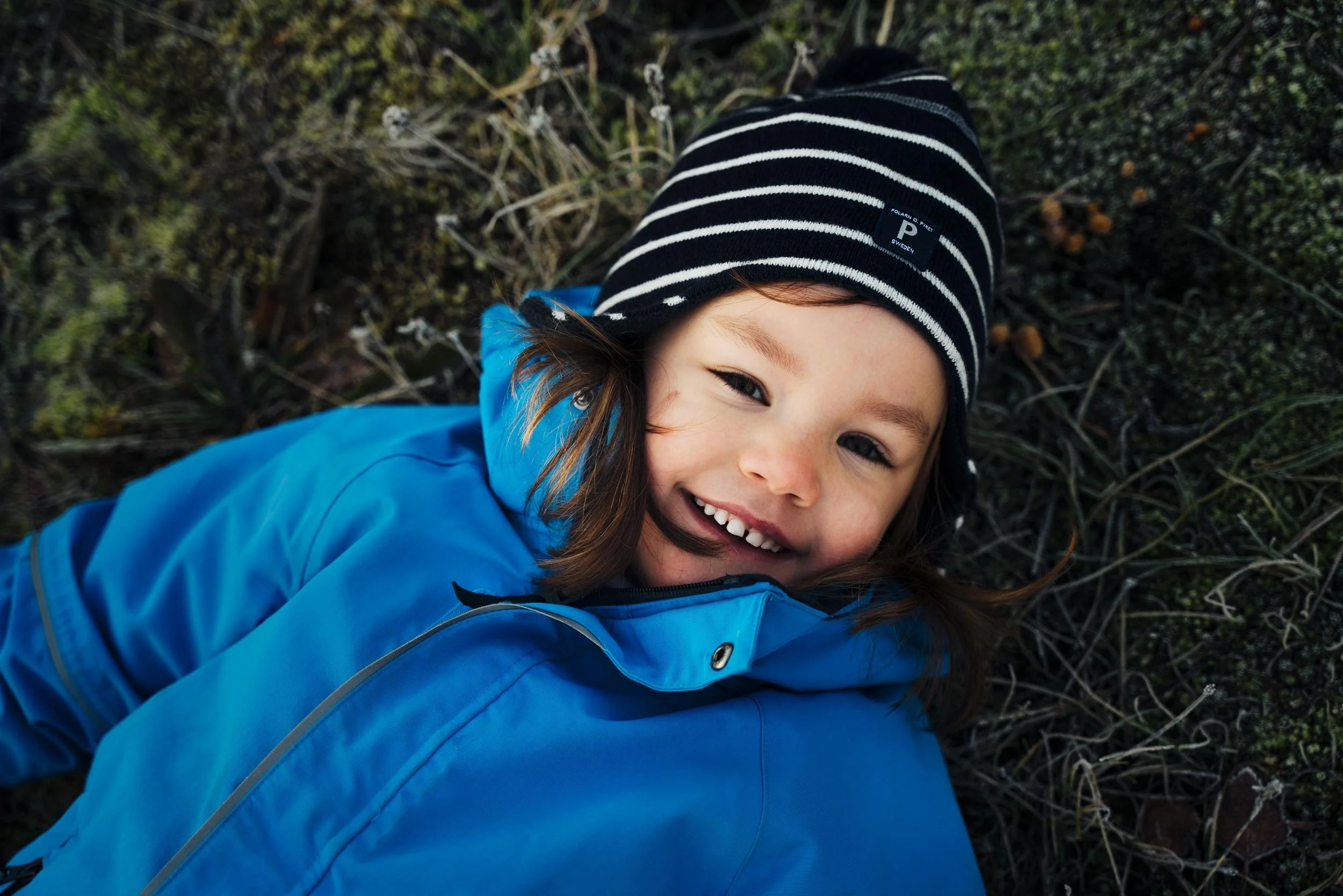 Young girl smiling outdoors, wearing a black and white striped beanie and a blue jacket, lying on the ground surrounded by grass and moss.