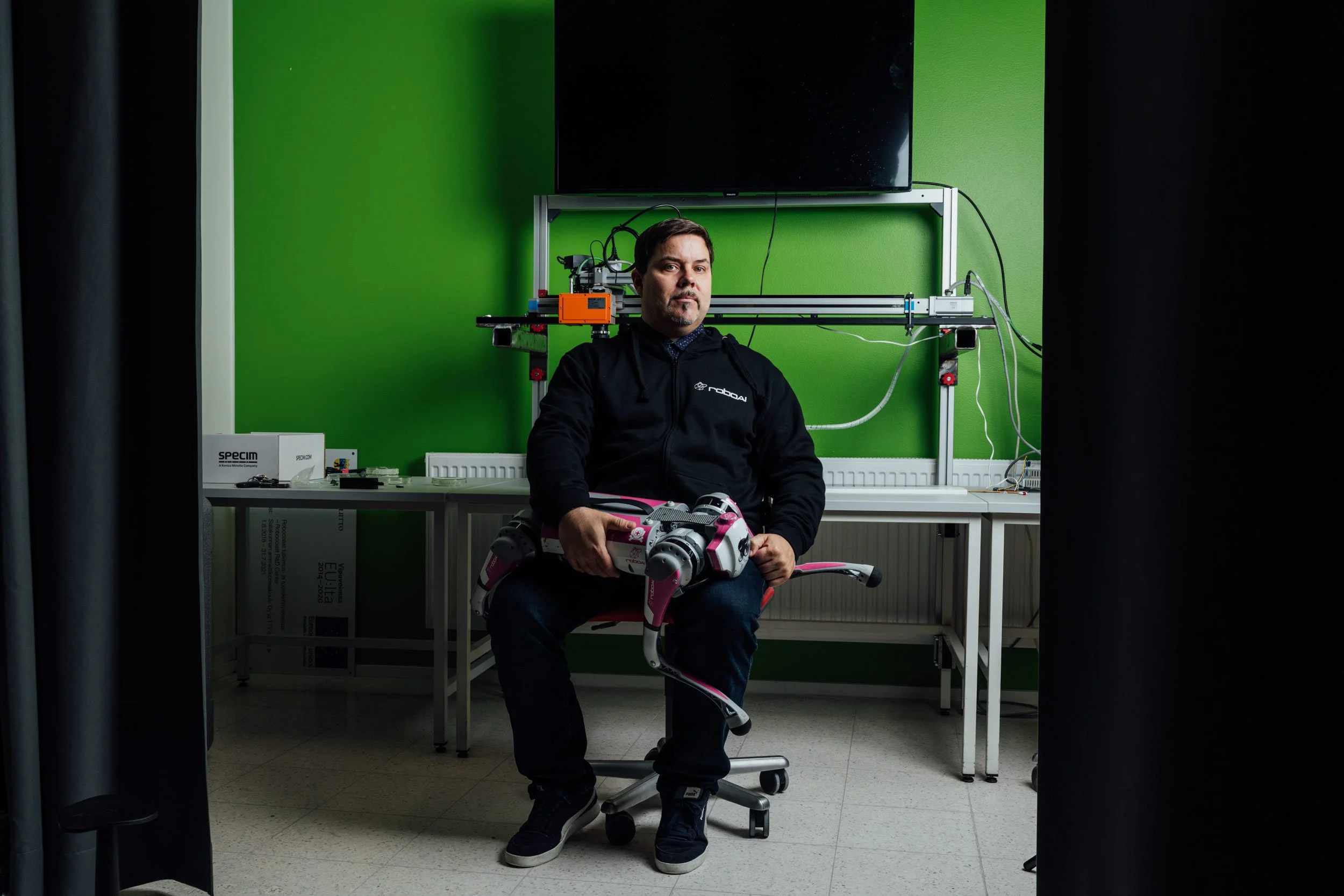 A man sitting in front of a green wall, holding a white and pink robotic dog, with a large TV and technical equipment behind him.