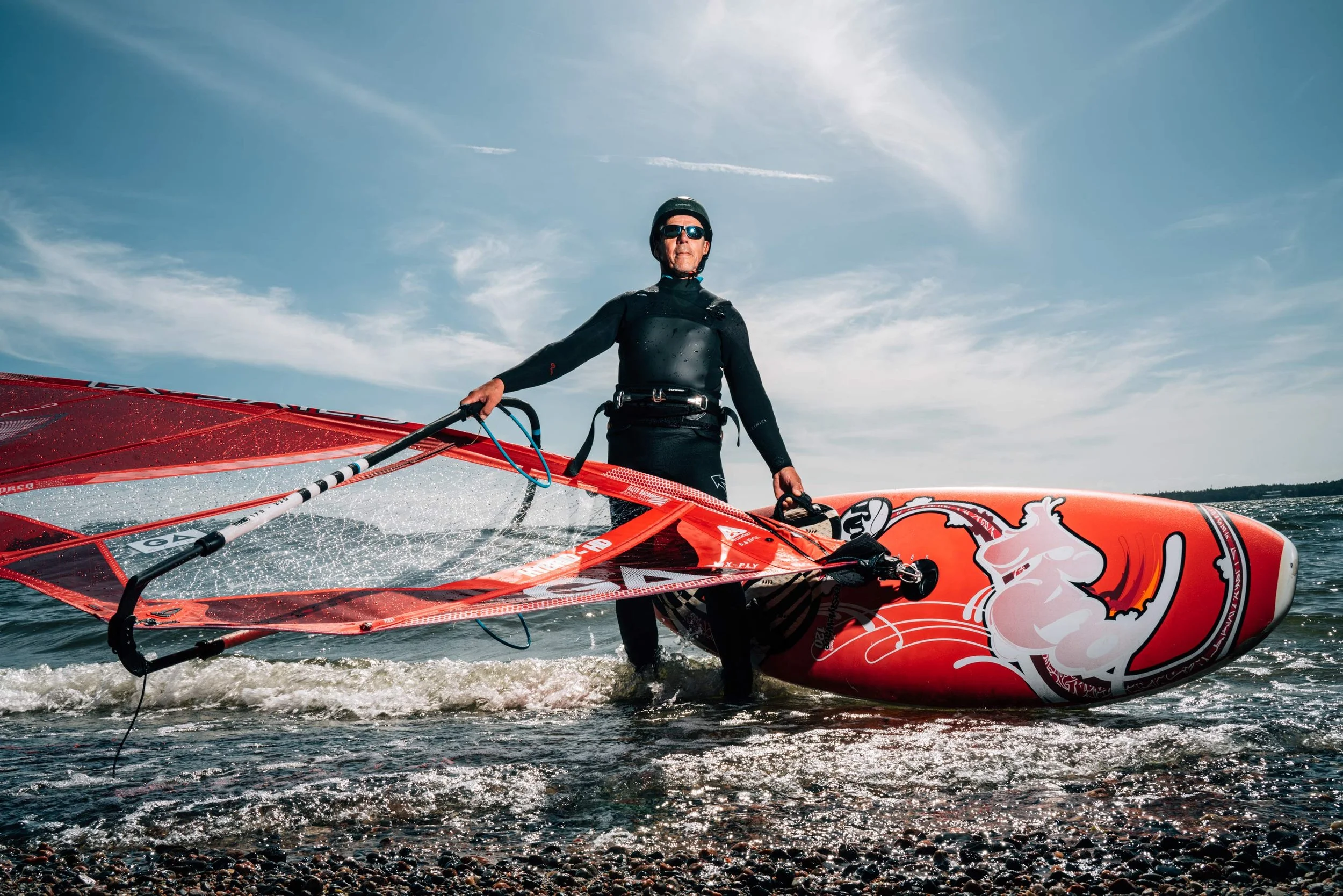 A man wearing sunglasses, a helmet, and a wetsuit standing in the water, holding a colorful windsurfing sail and board, with a partly cloudy sky overhead.