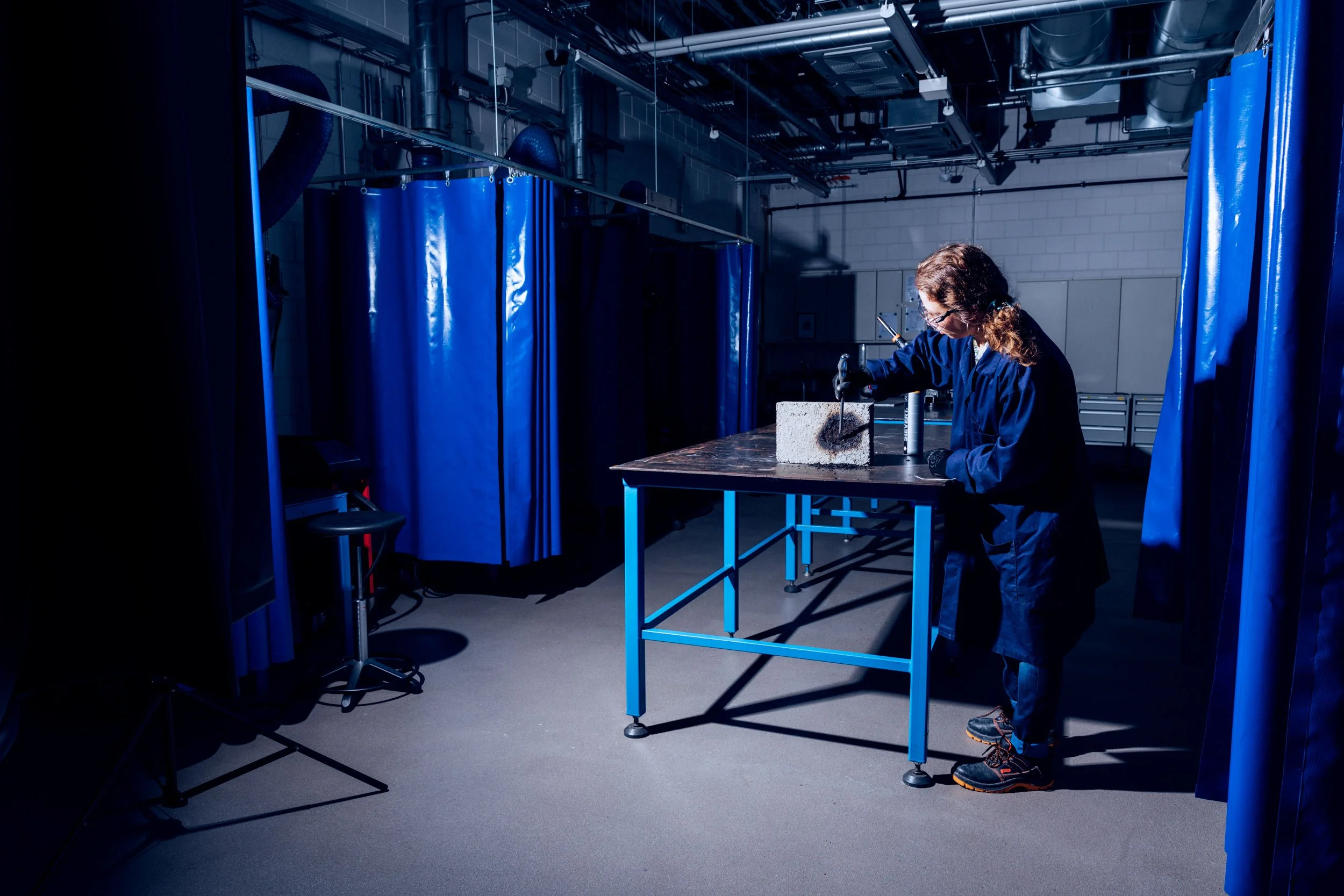 A person in safety gear working on a concrete block in a laboratory or industrial setting, with blue curtains and industrial pipes overhead.