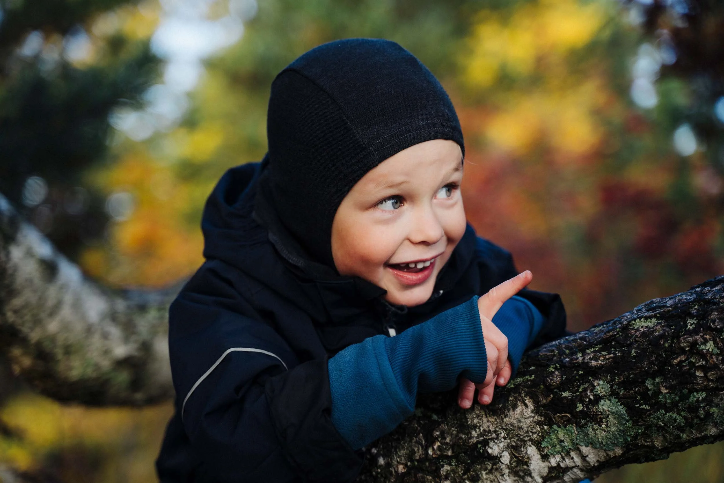 A young boy with a black hat and blue jacket smiling outdoors, touching a tree branch with a background of blurred autumn foliage.