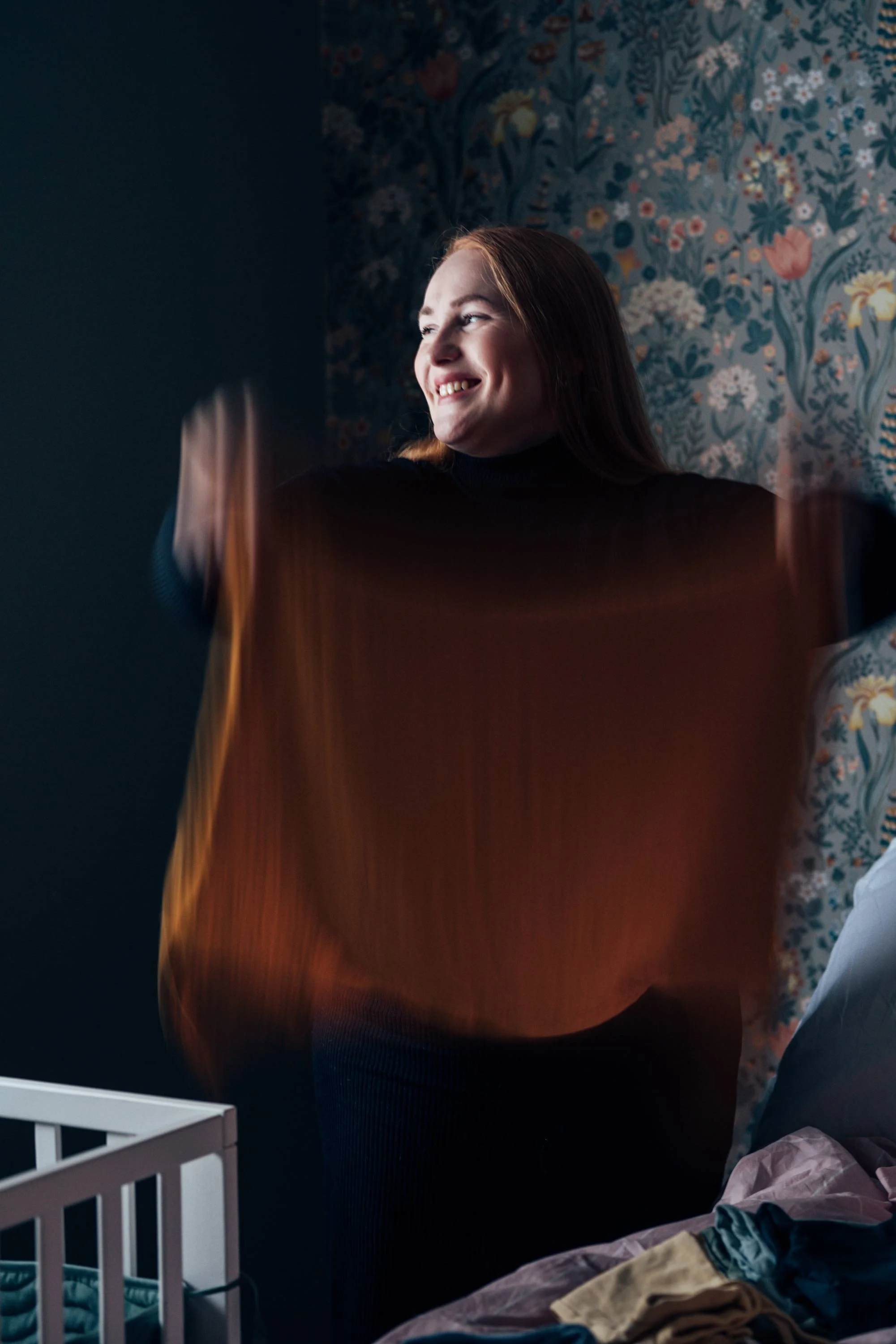 A smiling woman with reddish-brown hair, wearing a black turtleneck, standing in a room with blue floral wallpaper, next to a bed with clothes and a laundry basket.
