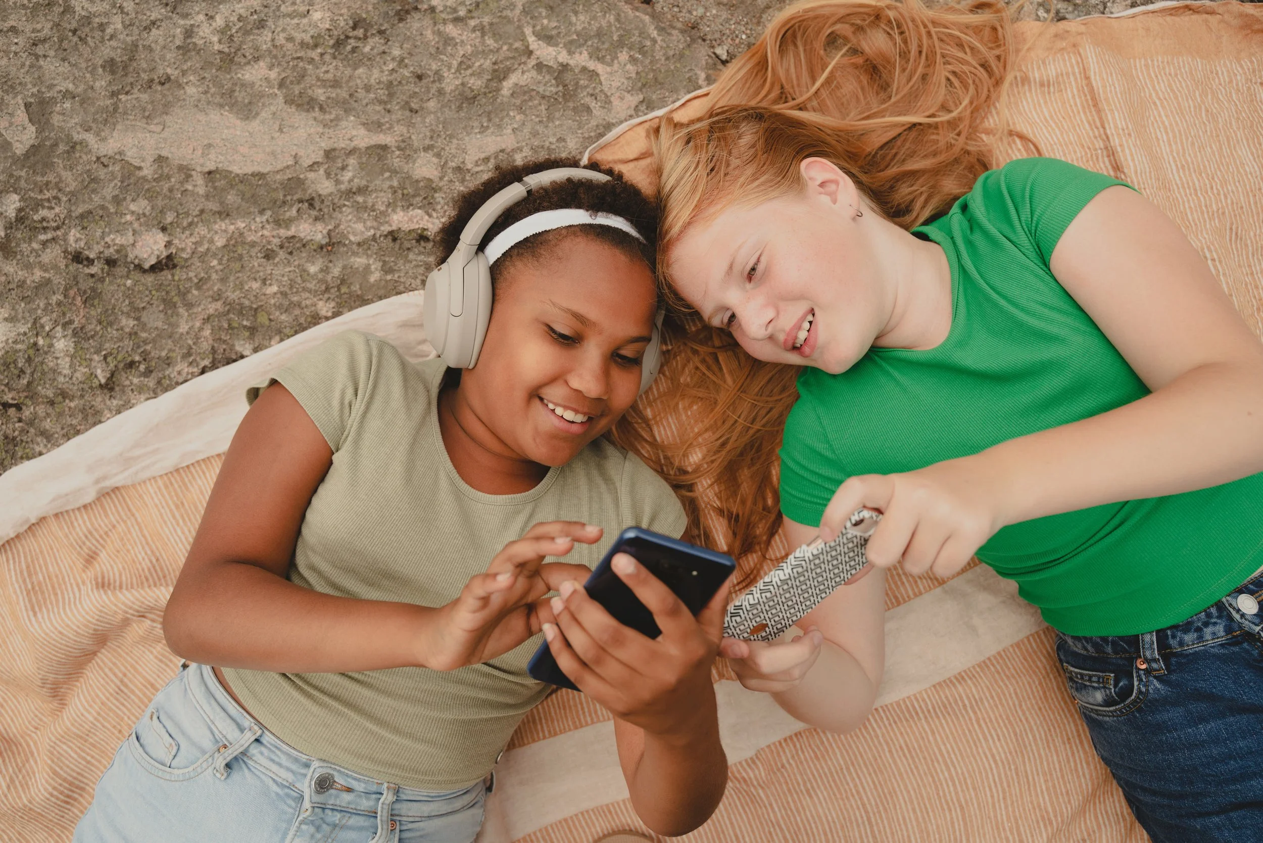 Two young girls lying on a striped blanket on the ground, looking at a smartphone together. One girl is wearing a green shirt and the other is wearing a beige shirt, with one girl also wearing headphones.