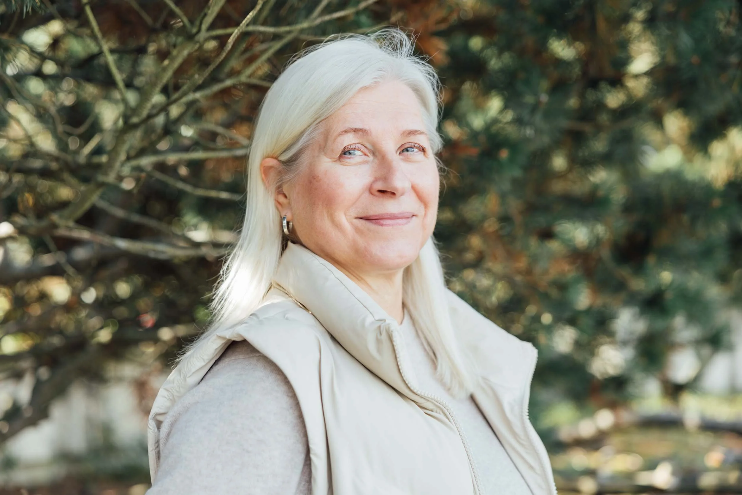 A woman with long white hair smiling outdoors, with trees and foliage in the background.