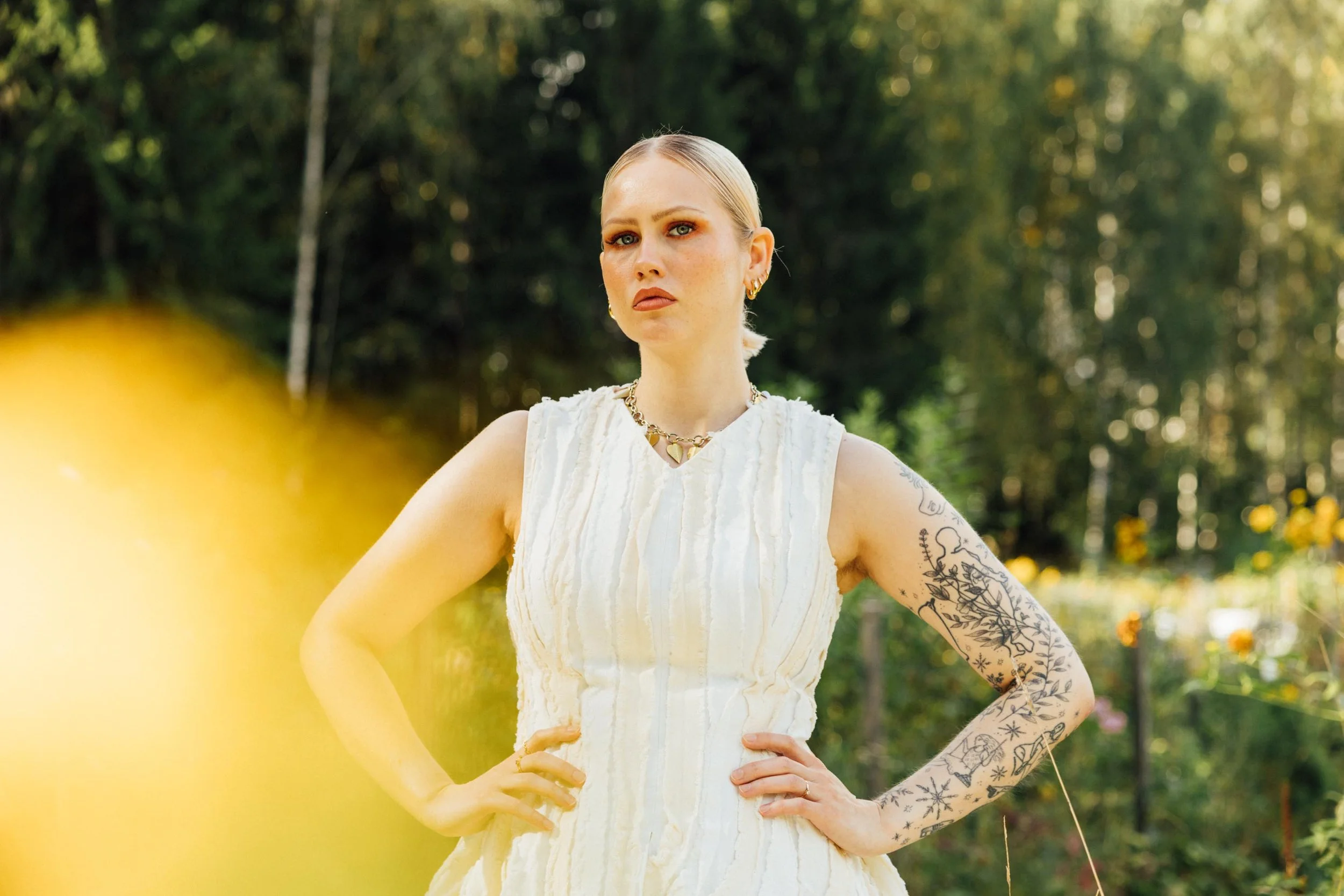 A woman with tattoos on her arms wearing a white sleeveless dress stands outdoors with a background of trees and sunlight.