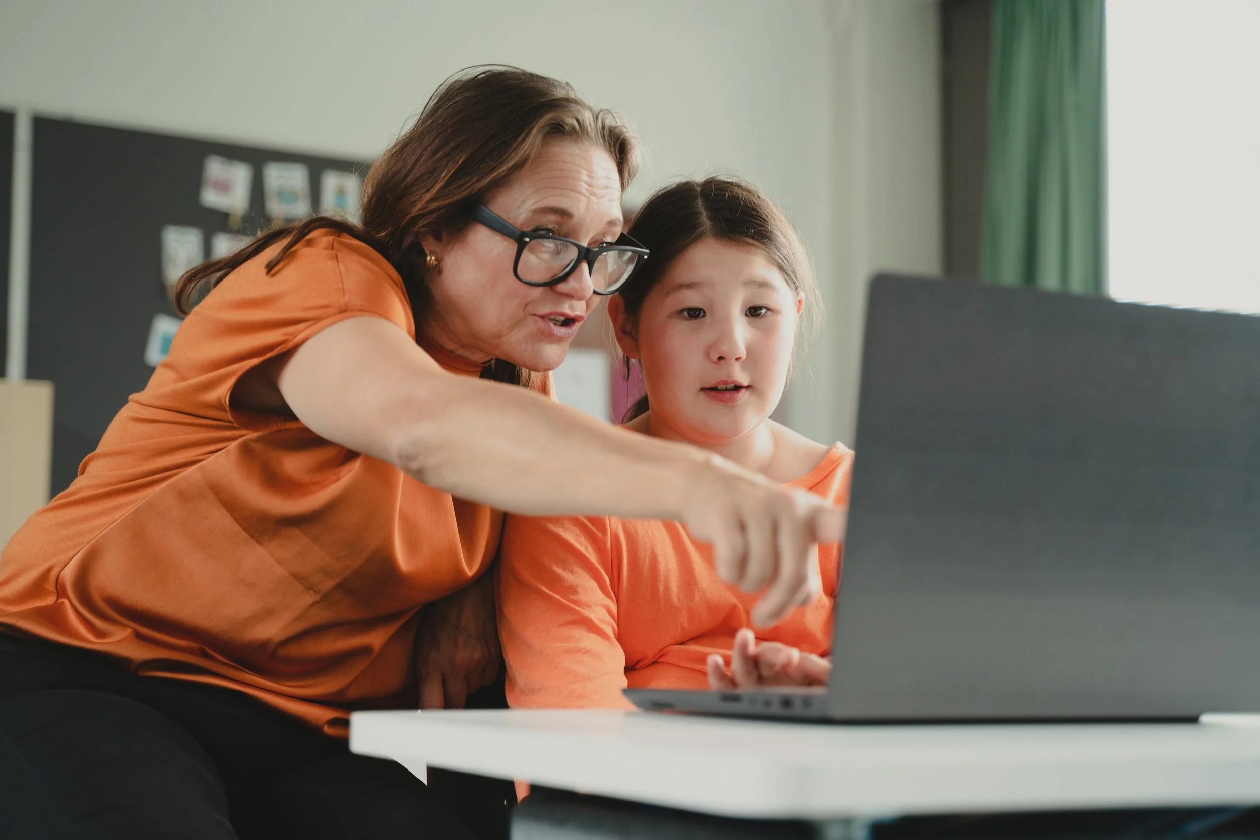 An adult woman and young girl sitting at a desk, looking at a laptop screen together, both wearing matching orange shirts, in a classroom setting.