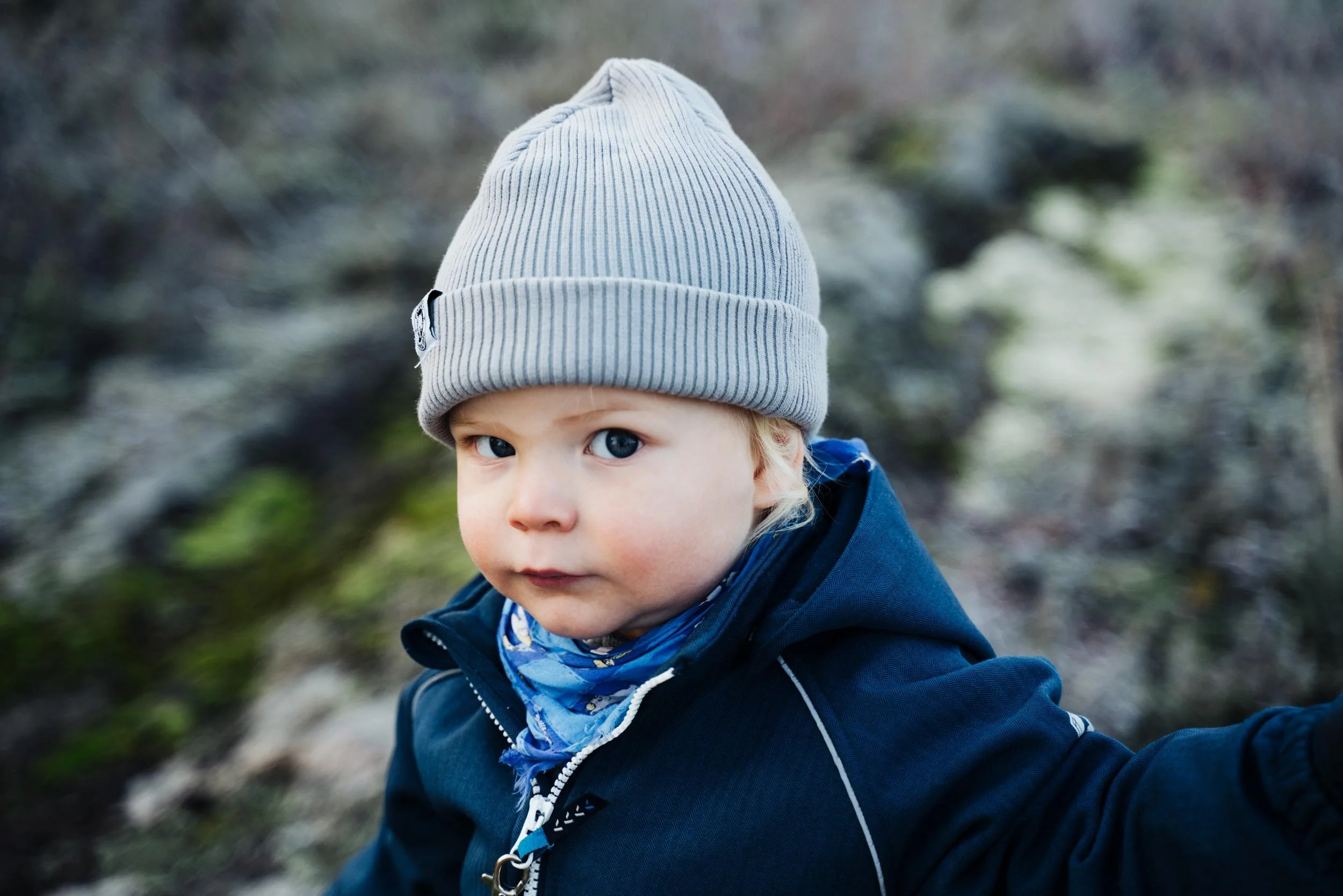 A young child with blue eyes wearing a gray knit beanie and a dark blue jacket outdoors.