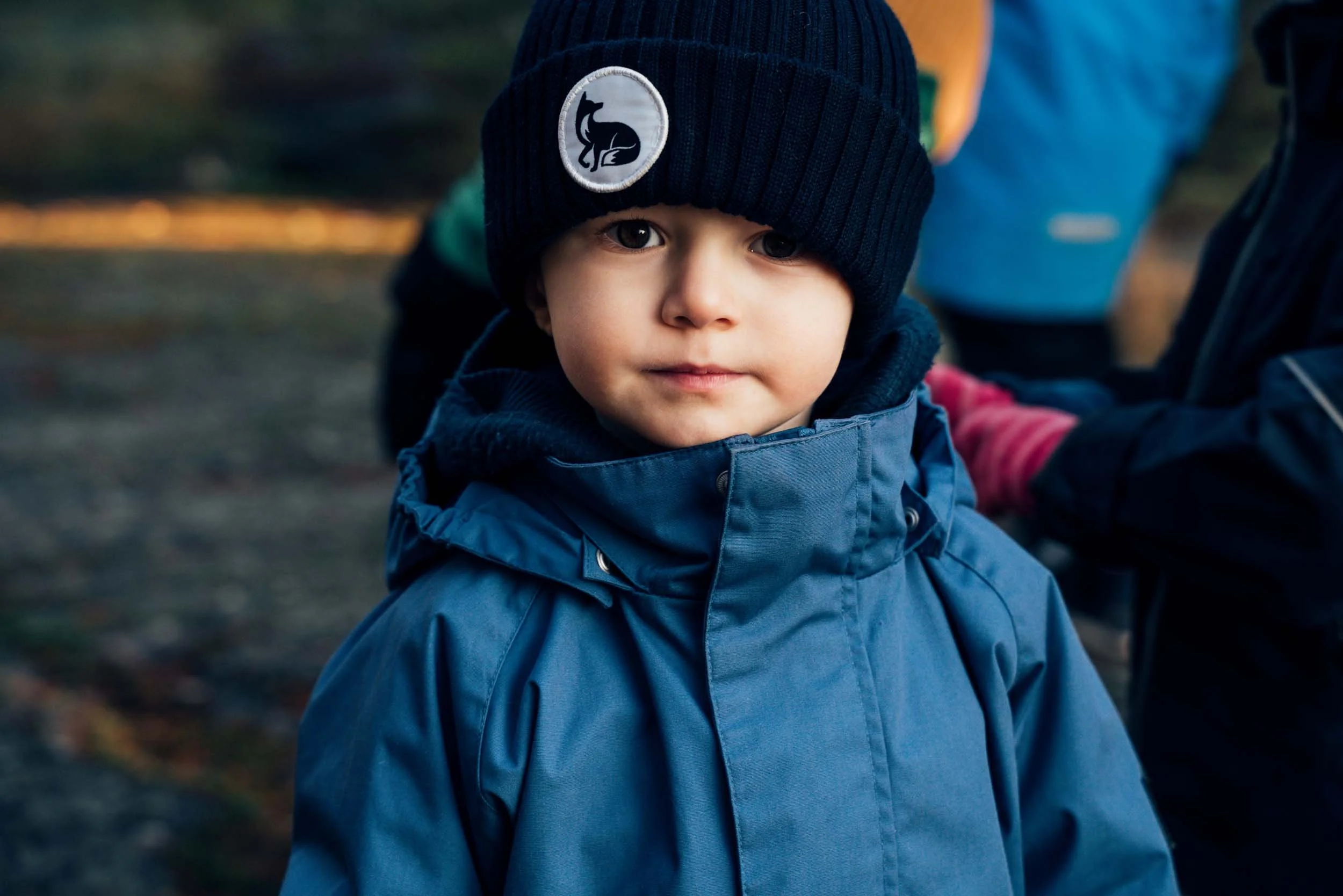 Close-up of a young boy wearing a dark blue knit hat with a logo of a sitting black dog, a blue jacket, and a black hoodie, outdoors with blurred background of trees and ground.