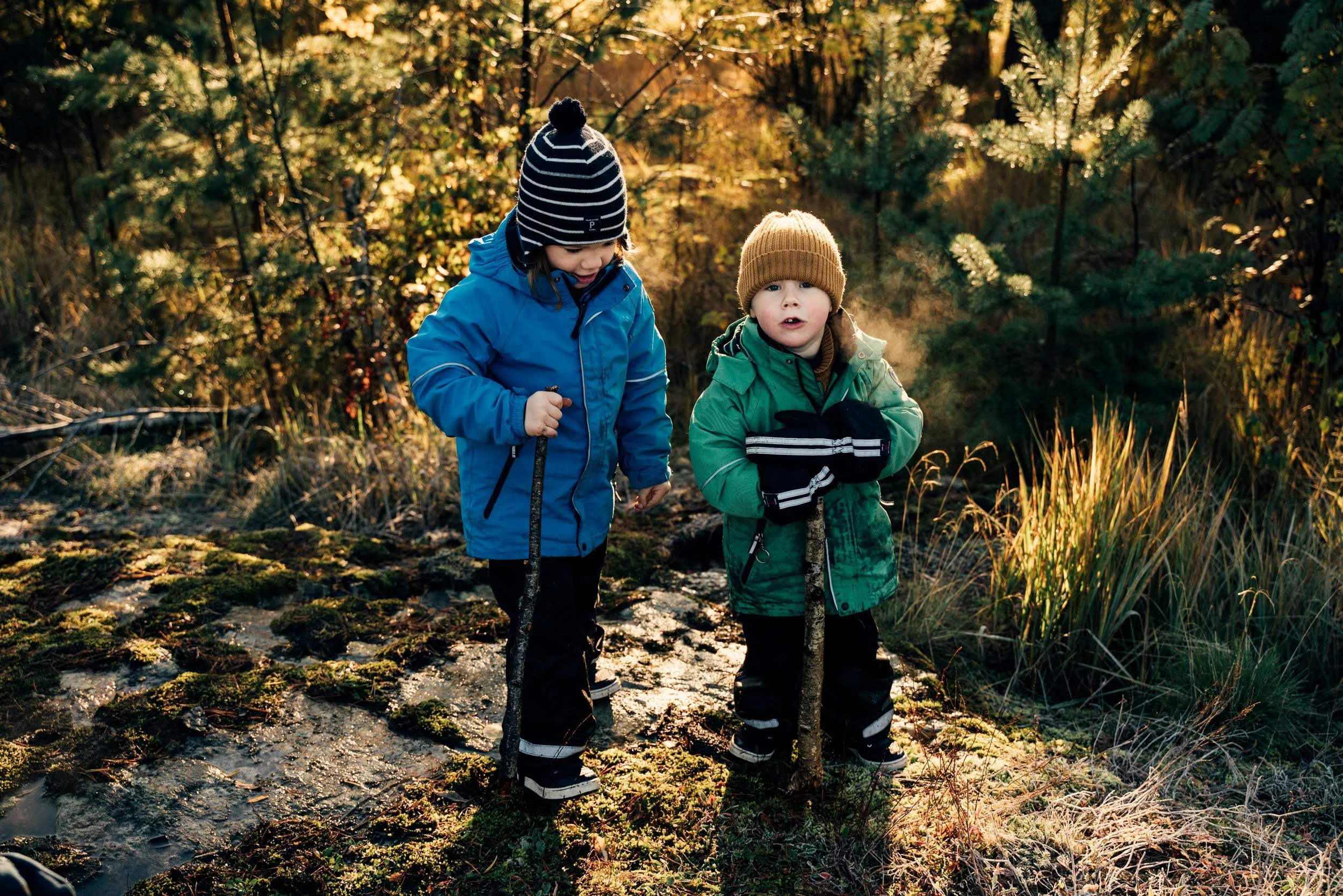 Two children dressed in winter jackets, hats, and gloves walking through a forest with sunlight filtering through the trees, holding stick-like objects.