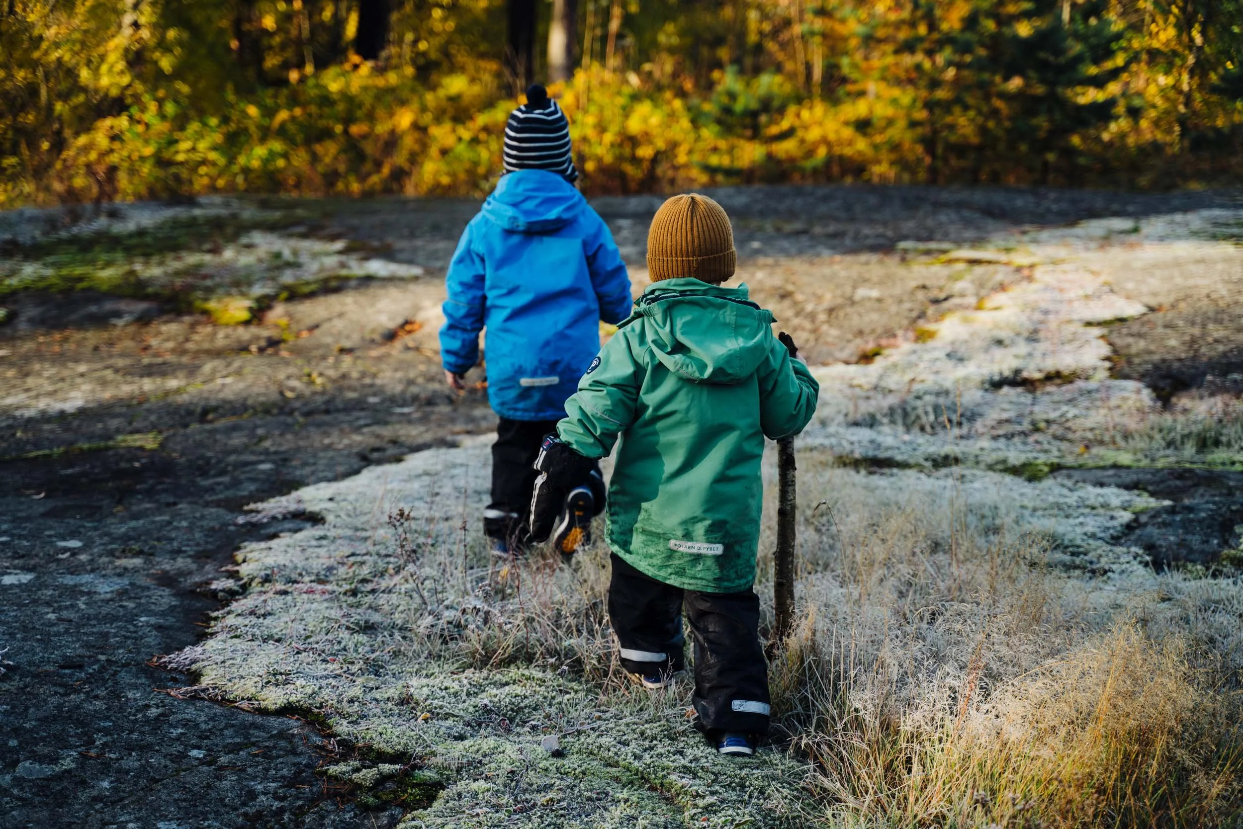 Two children walking outdoors on a rocky trail in fall, wearing jackets and hats, with trees displaying autumn foliage in the background.
