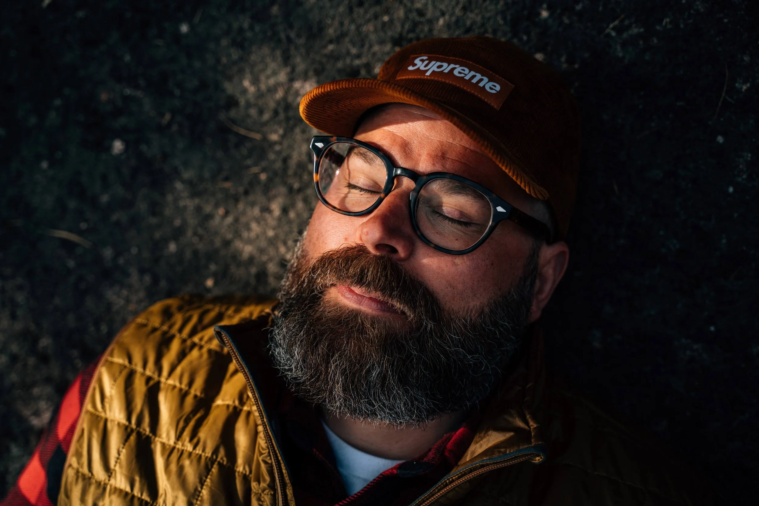 A bearded man with glasses and closed eyes, wearing a brown Supreme hat and a yellow and red jacket, lying on the ground outdoors at night.